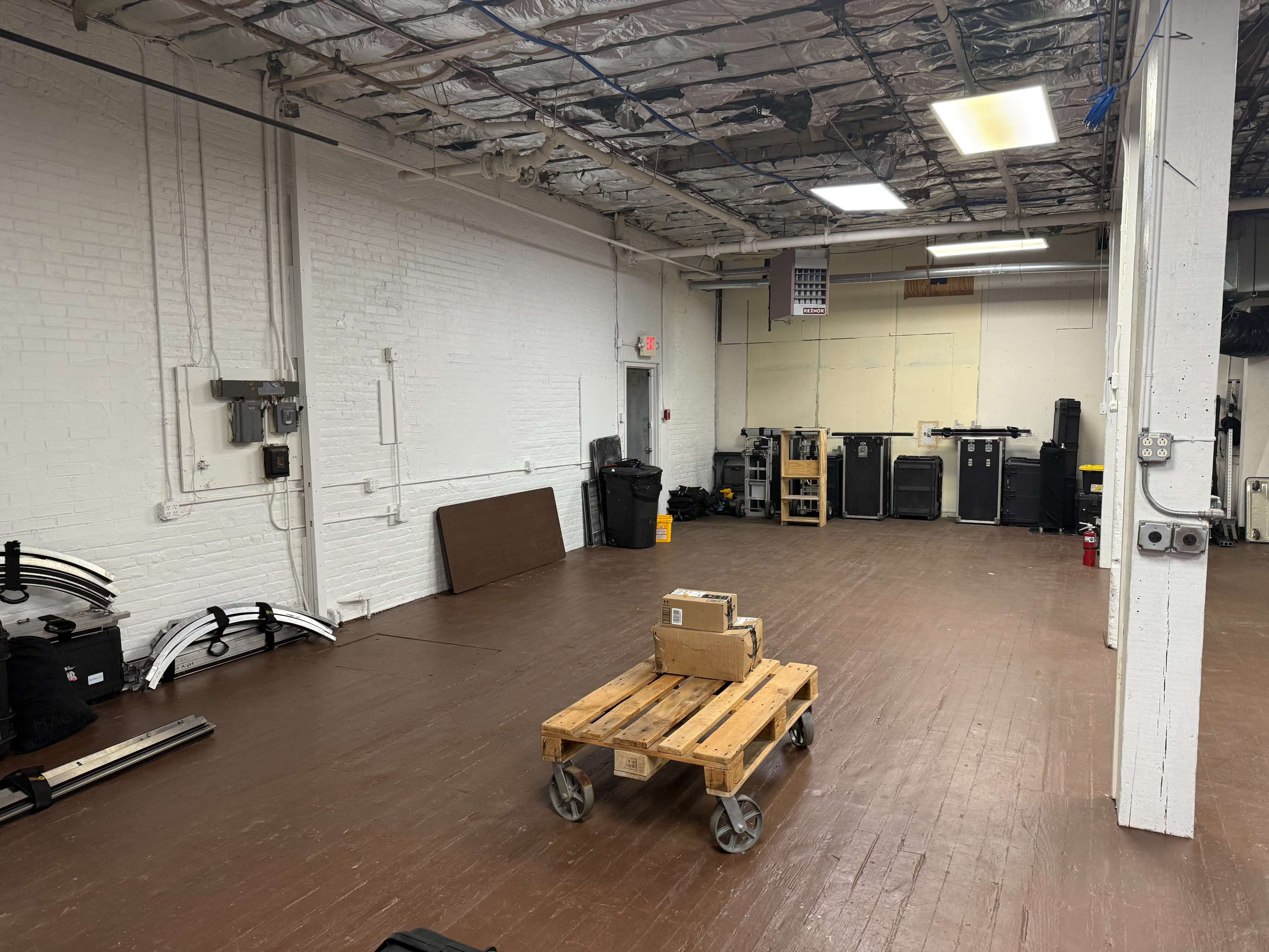 The image shows a large, empty industrial space with wooden flooring and several pieces of equipment and gear stored against the walls.