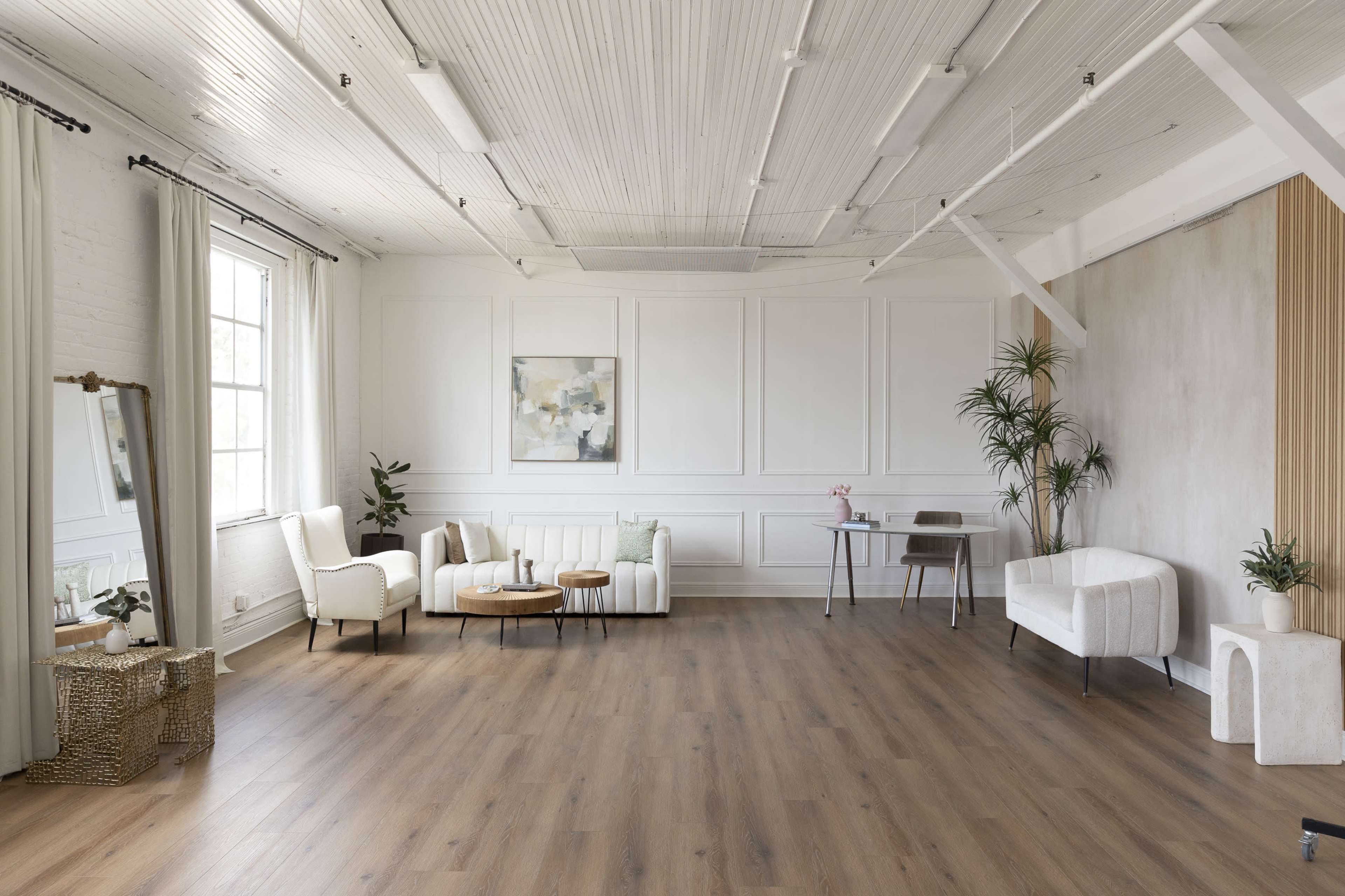 The image shows a spacious, well-lit interior featuring minimalist furniture arrangements, a large window, and decorative plants.