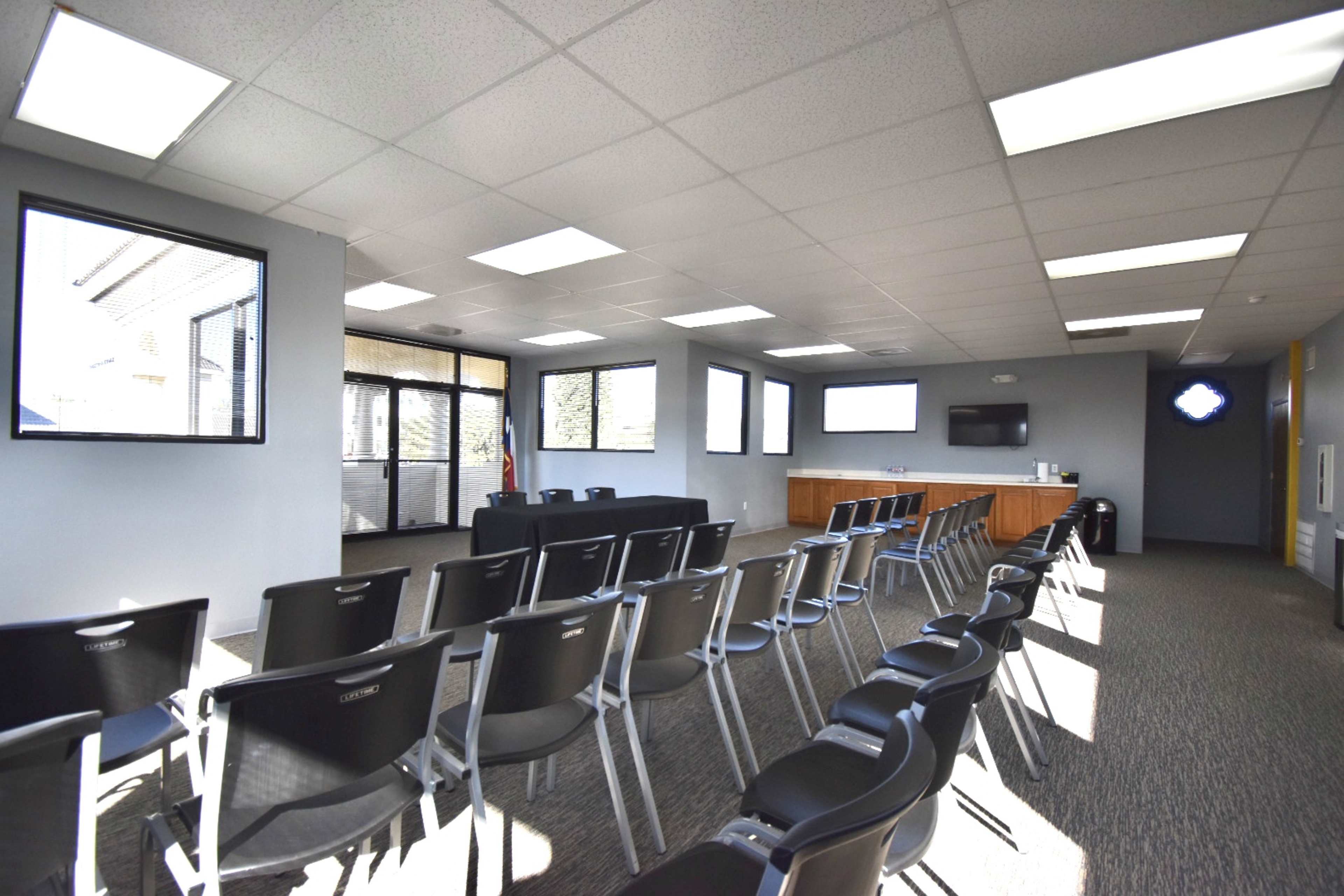 The image shows a spacious meeting room with rows of black chairs facing a presentation area, equipped with windows and a television screen.