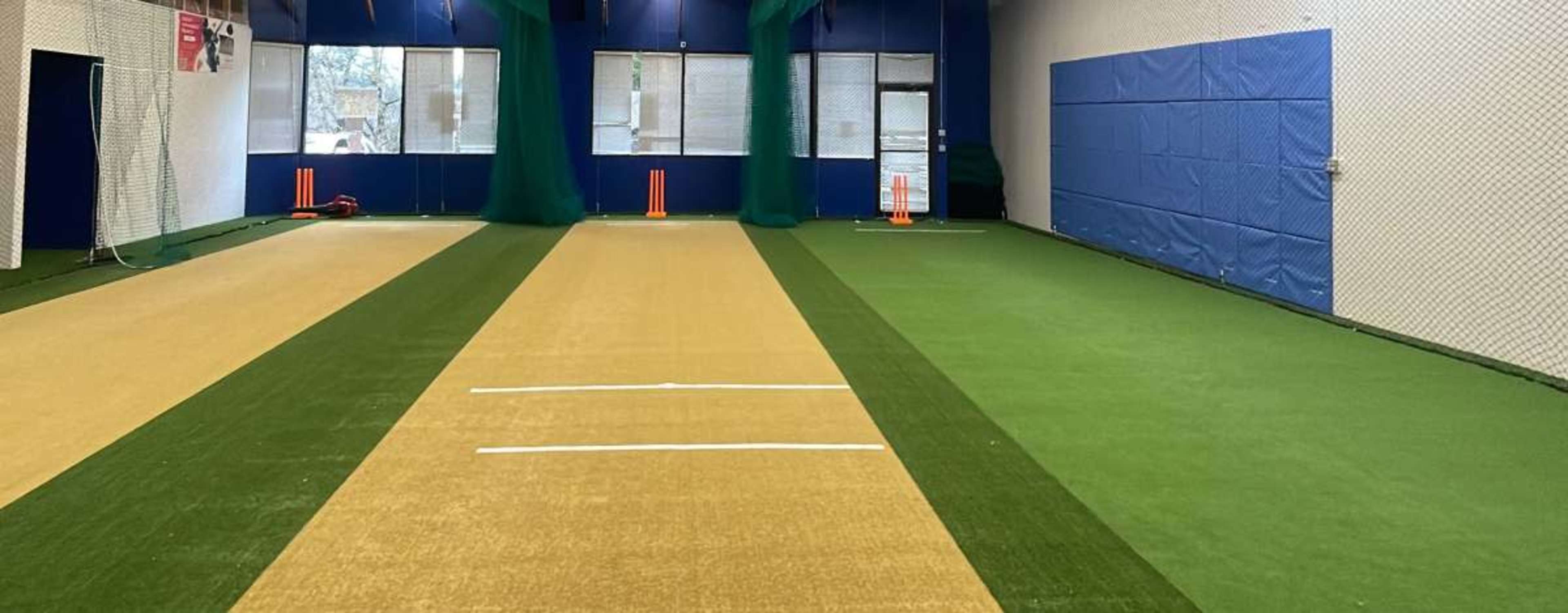 The image shows an indoor sports facility with two cricket practice lanes, each marked with white lines, surrounded by green turf and netting.