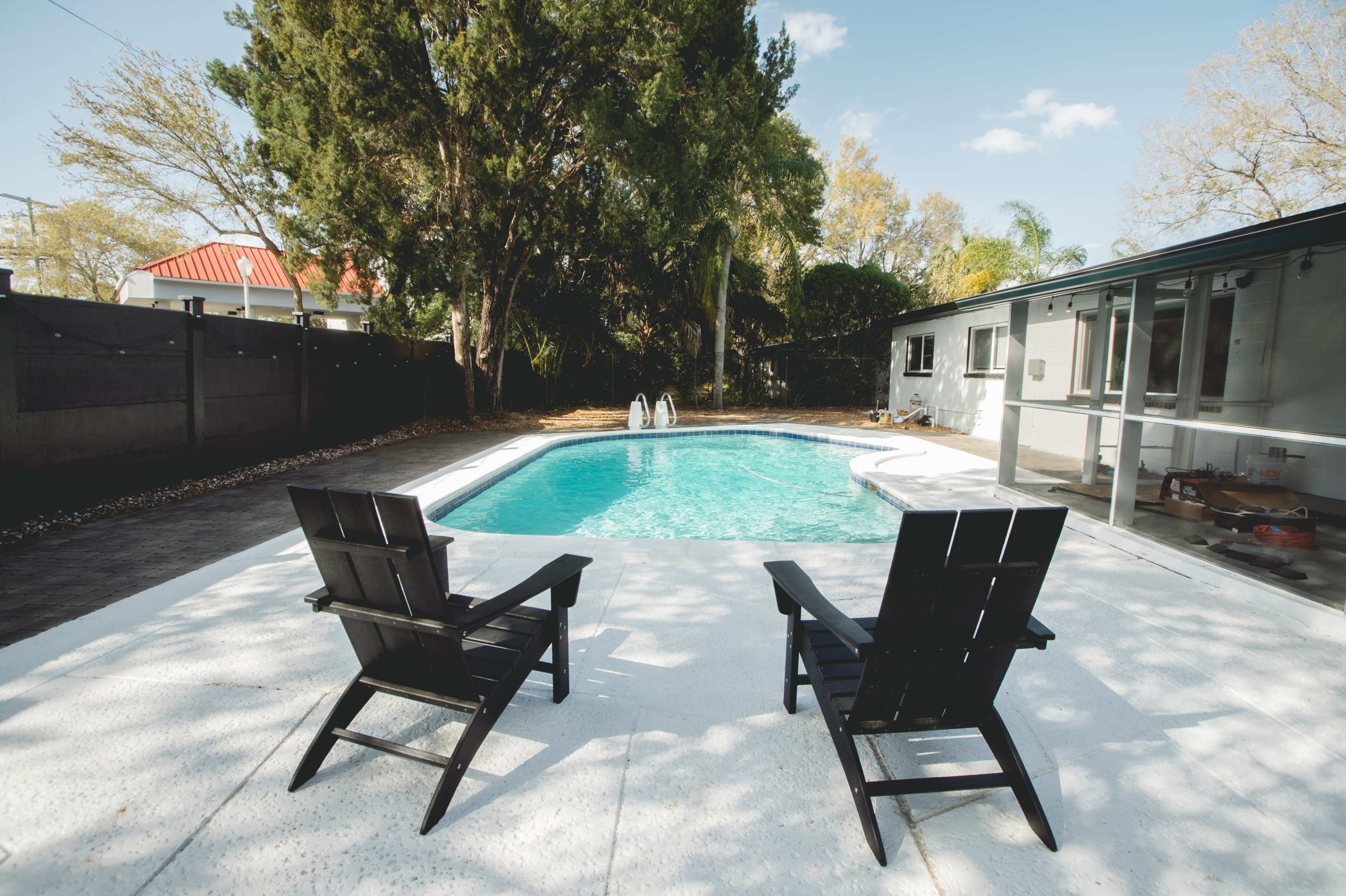 Two black lounge chairs face a swimming pool surrounded by a concrete deck and lush greenery.