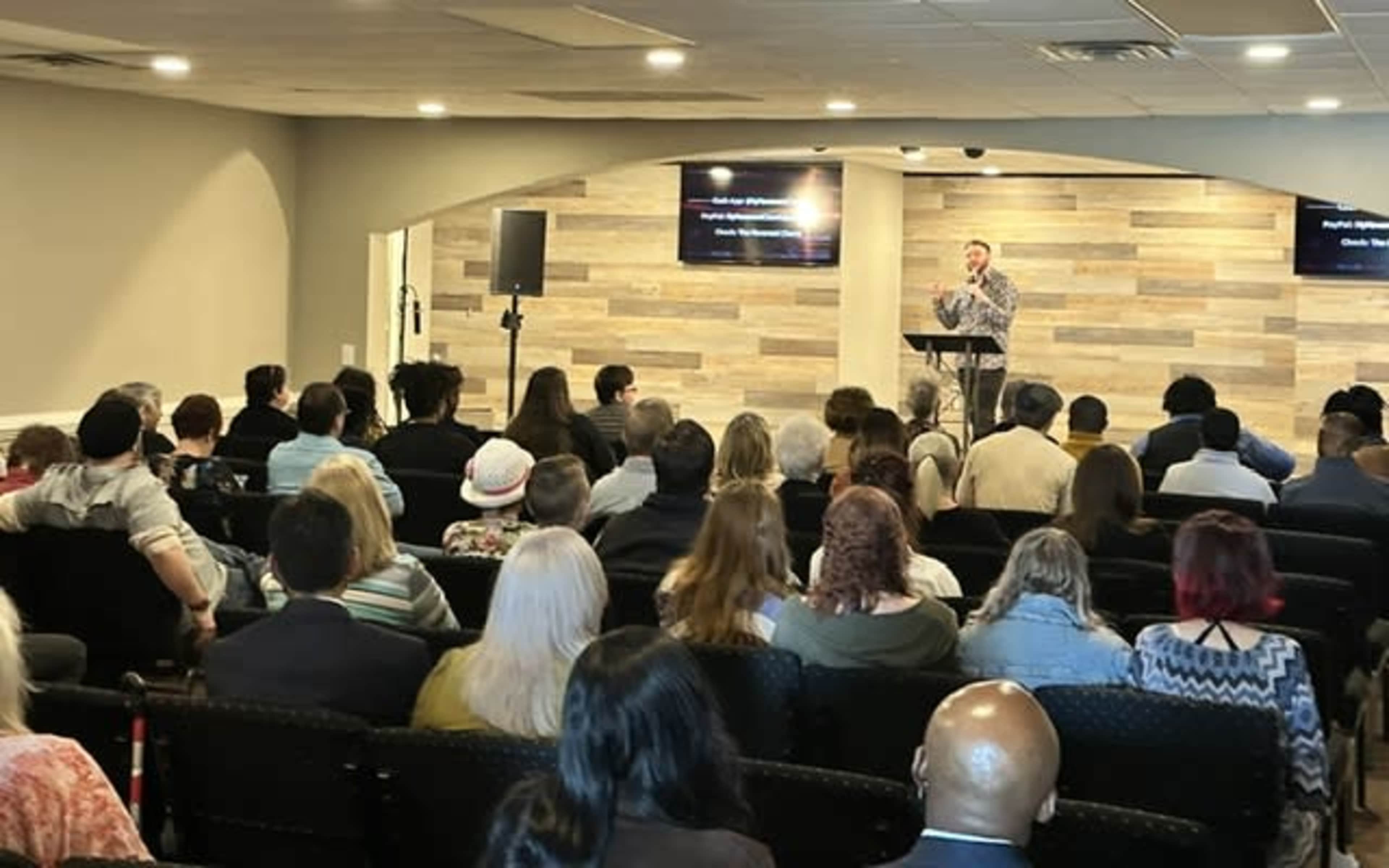 A speaker stands at a podium addressing a large audience seated in a spacious room.