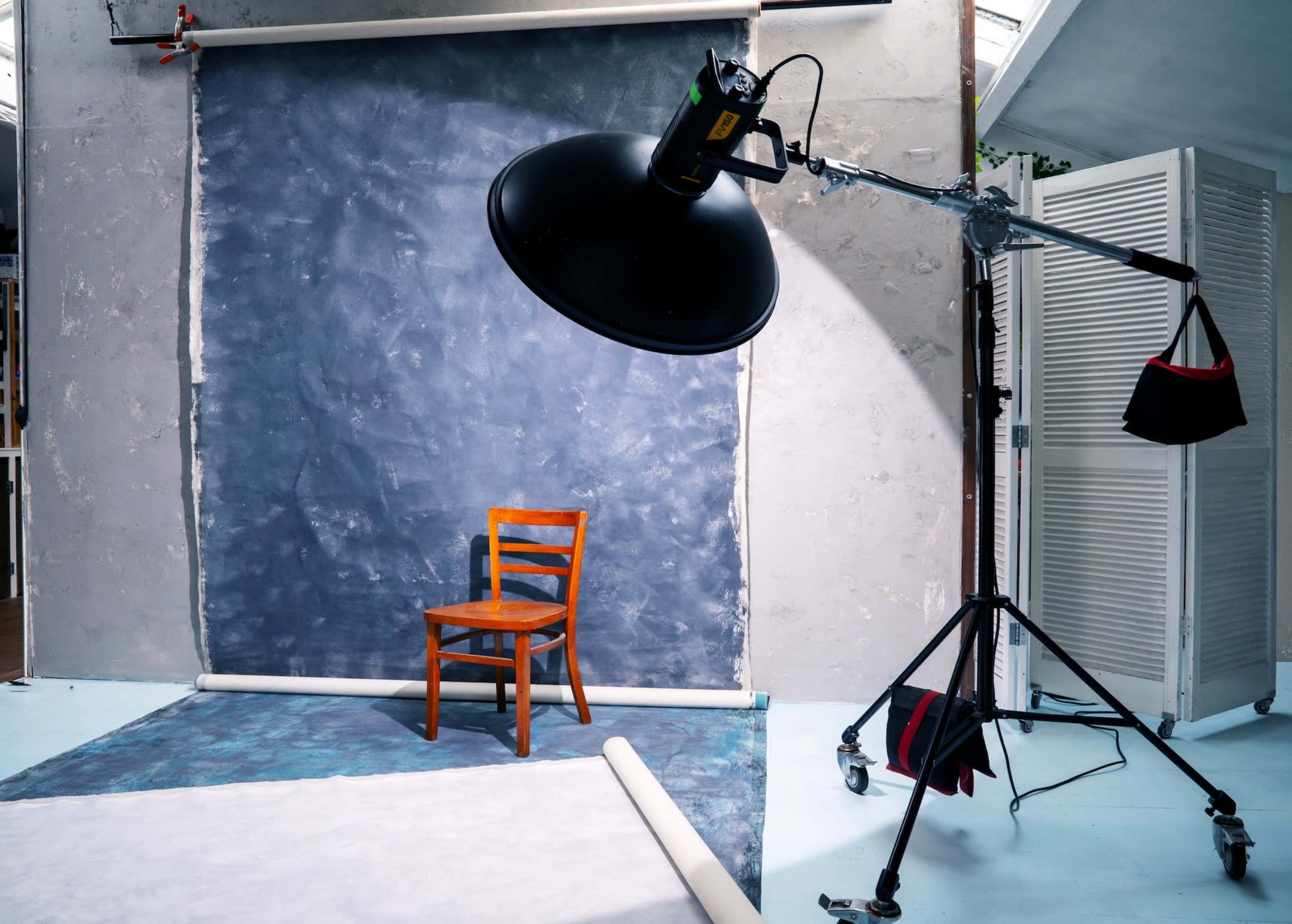 The image shows a photography studio with a wooden chair positioned in front of a blue backdrop under a large studio light.