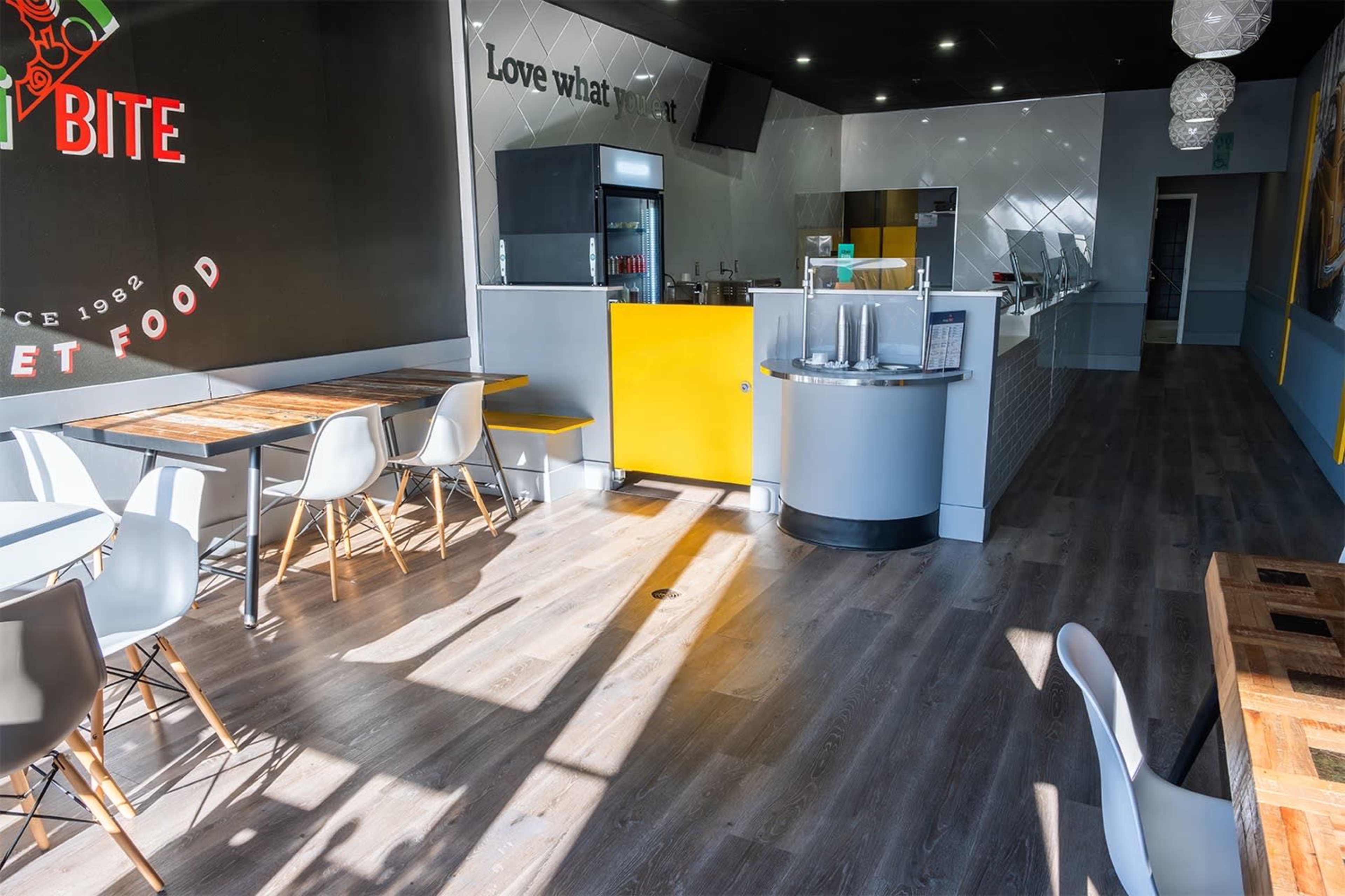 A modern, vacant restaurant interior with wooden tables and white chairs, featuring a service counter and a wall with motivational text.
