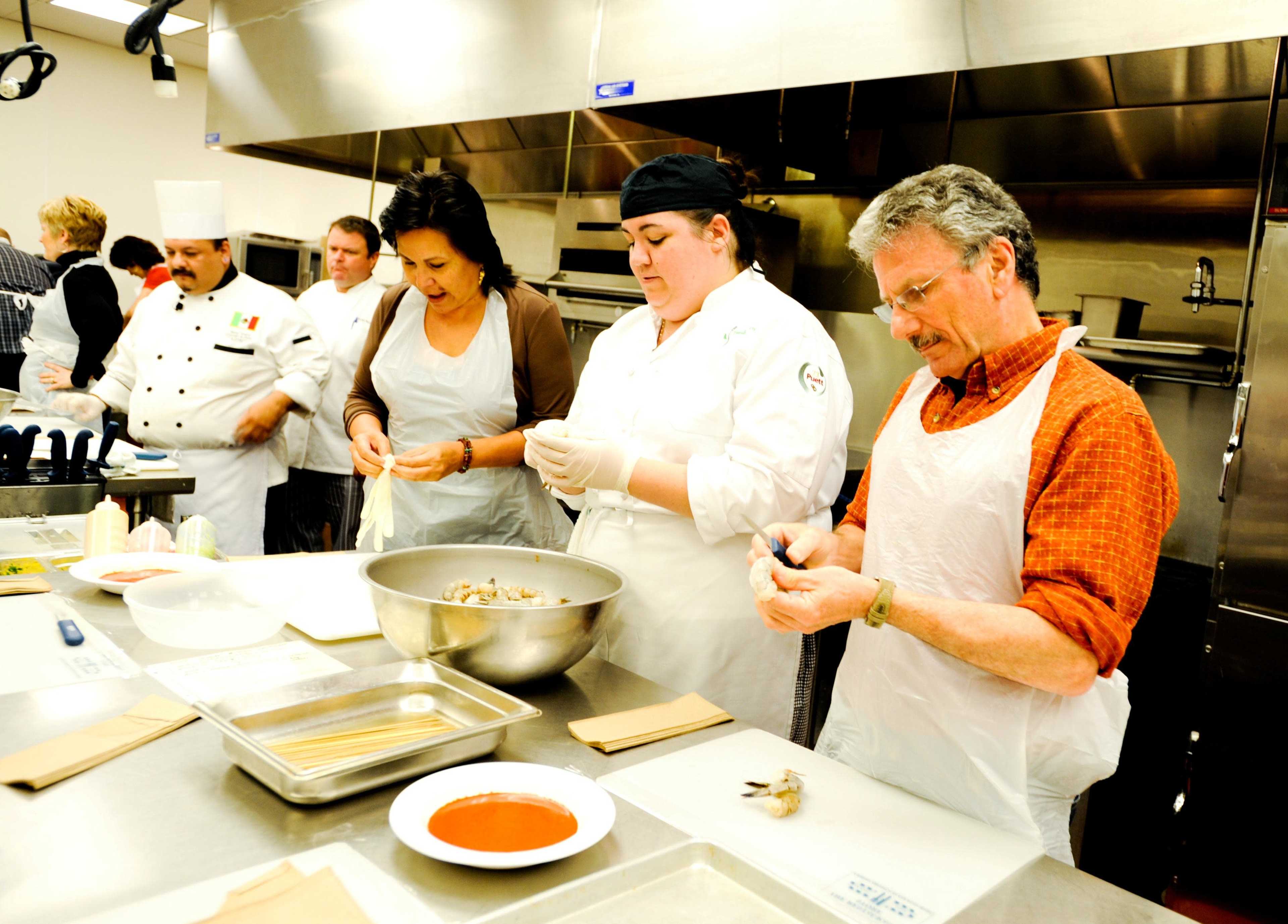 Several individuals are preparing food in a commercial kitchen, focused on cooking tasks while wearing aprons and gloves.