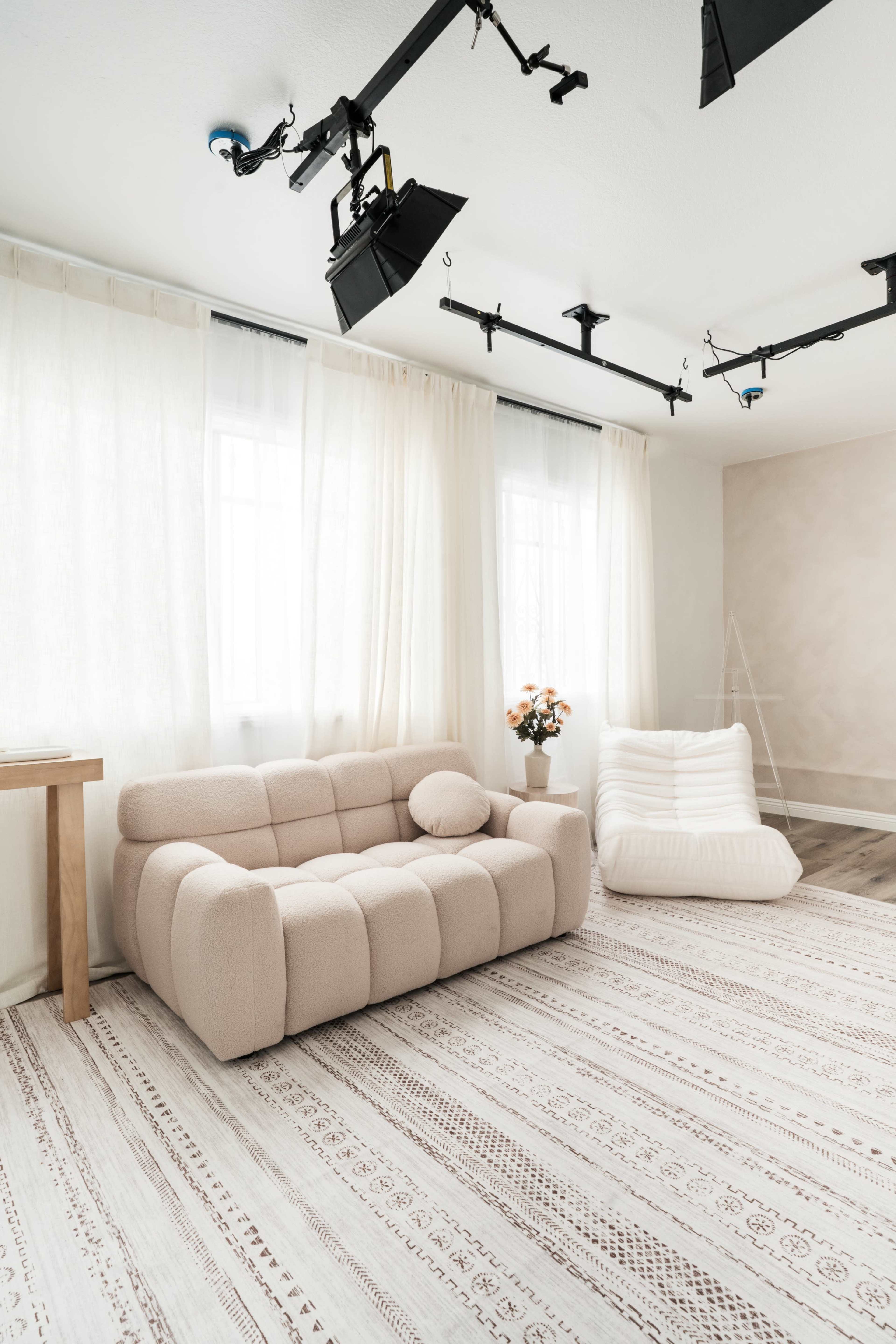 A minimalist living space features a beige upholstered sofa, a decorative flower arrangement, and soft textures on the floor and walls.
