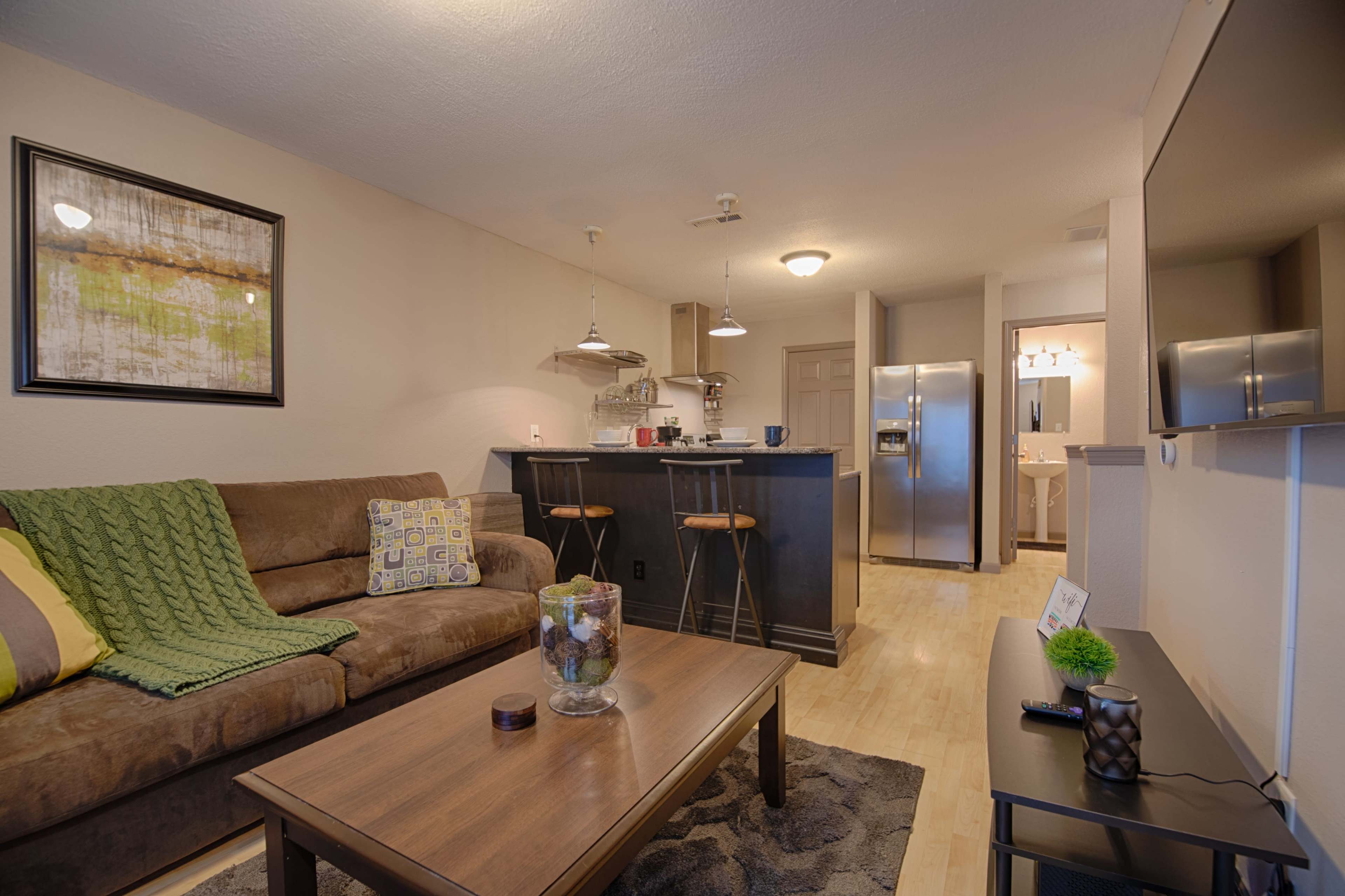 A living room features a brown sofa with a green throw, a wooden coffee table, and an adjoining kitchen area with a bar counter and stainless steel appliances.