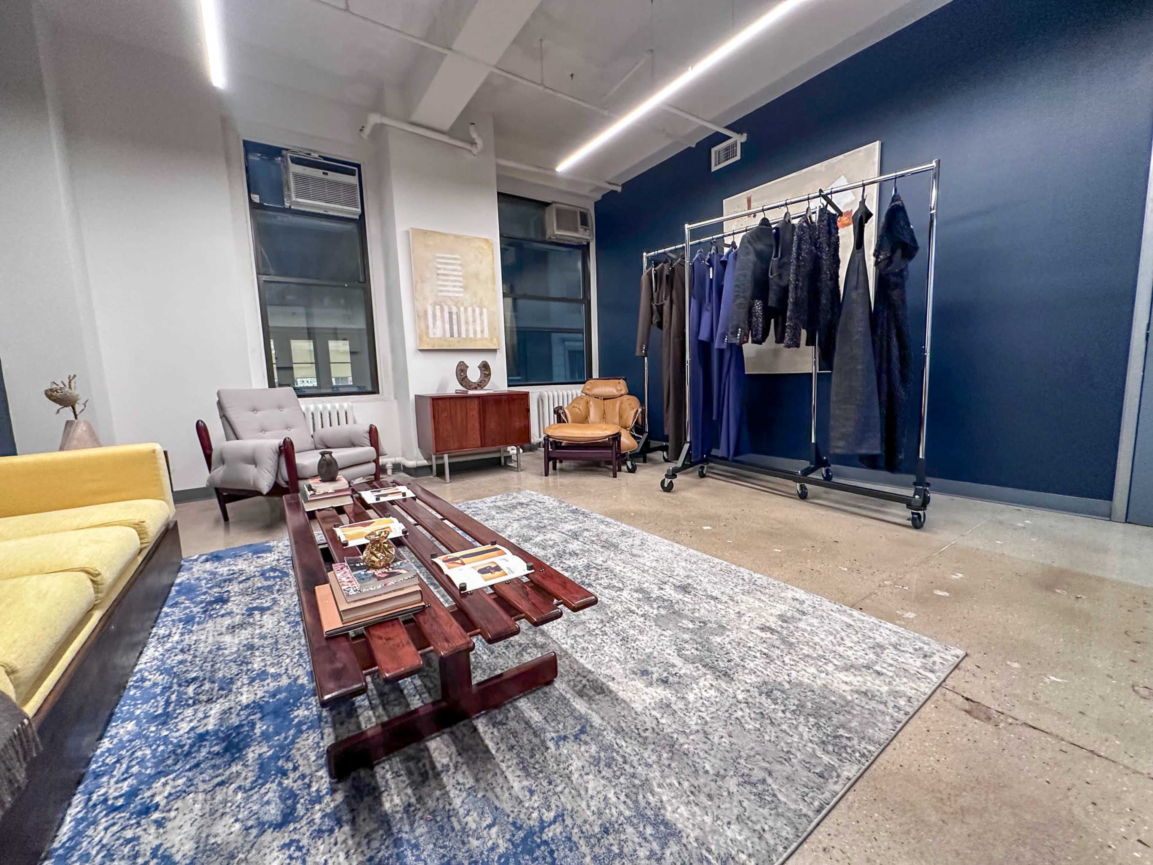 The image shows a modern interior space with a yellow sofa, a gray armchair, a wooden coffee table, a clothing rack with garments, and a blue accent wall.