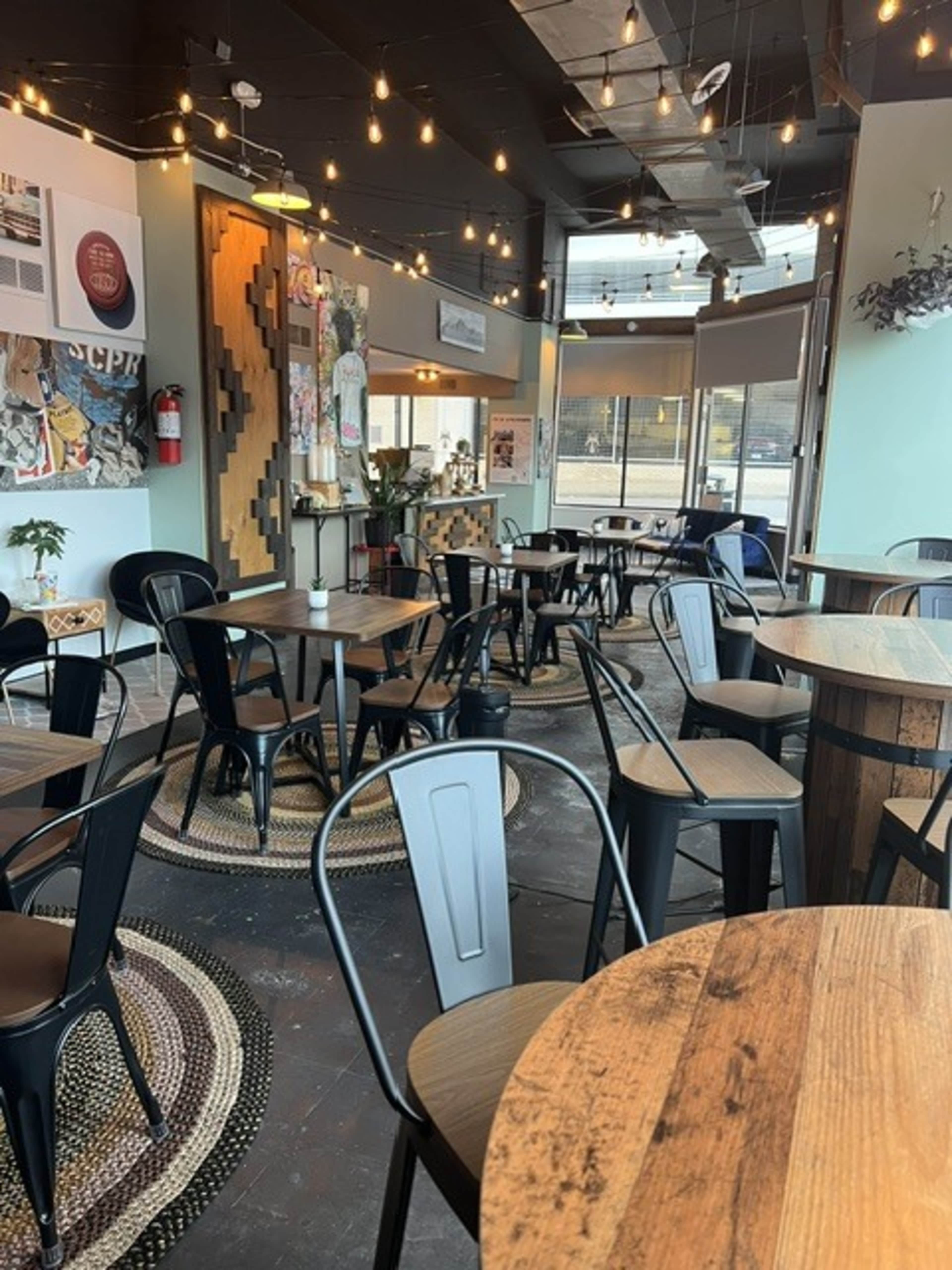 The image shows a cozy café interior with wooden tables, black chairs, and decorative lighting.