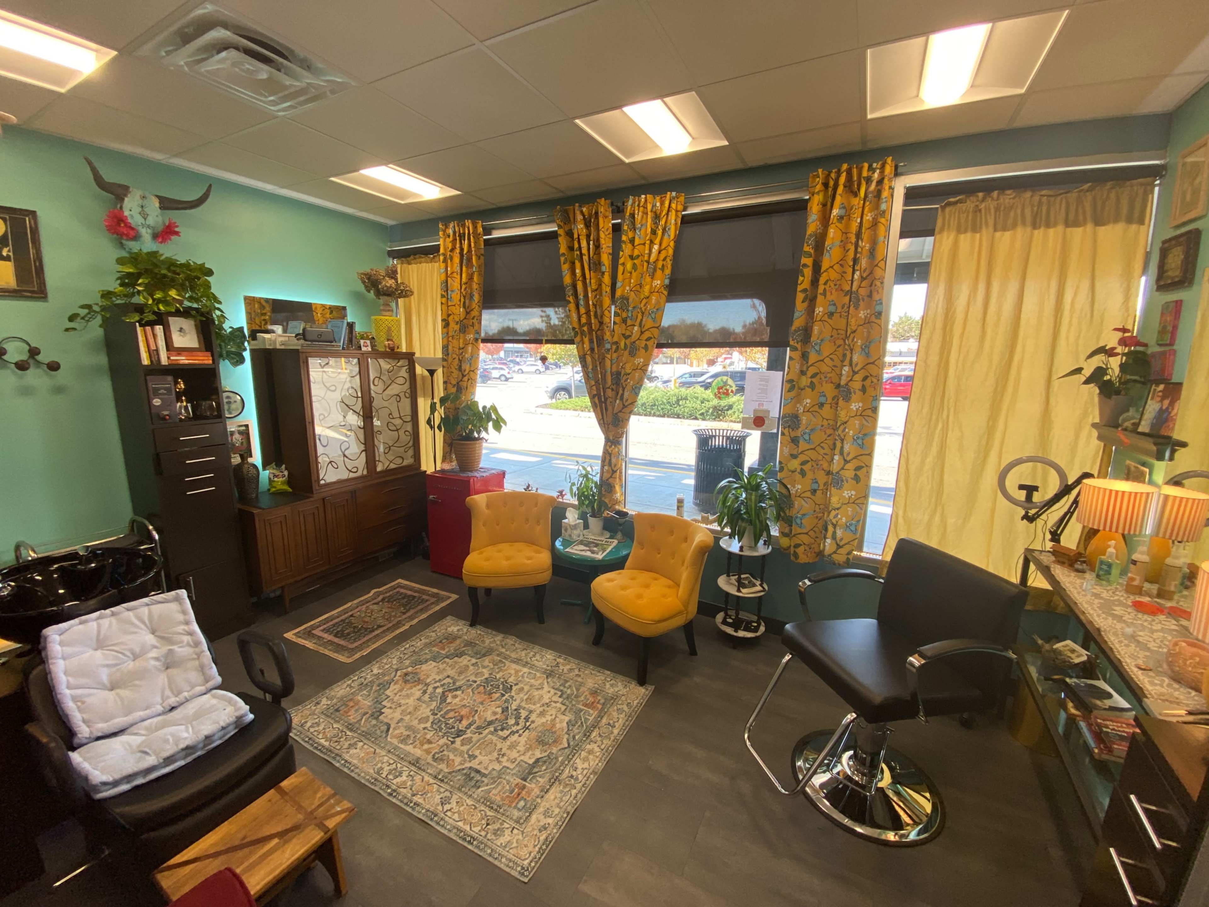 The image shows a brightly colored hair salon interior with yellow chairs, a small rug, a styling chair, and large windows covered with patterned curtains.