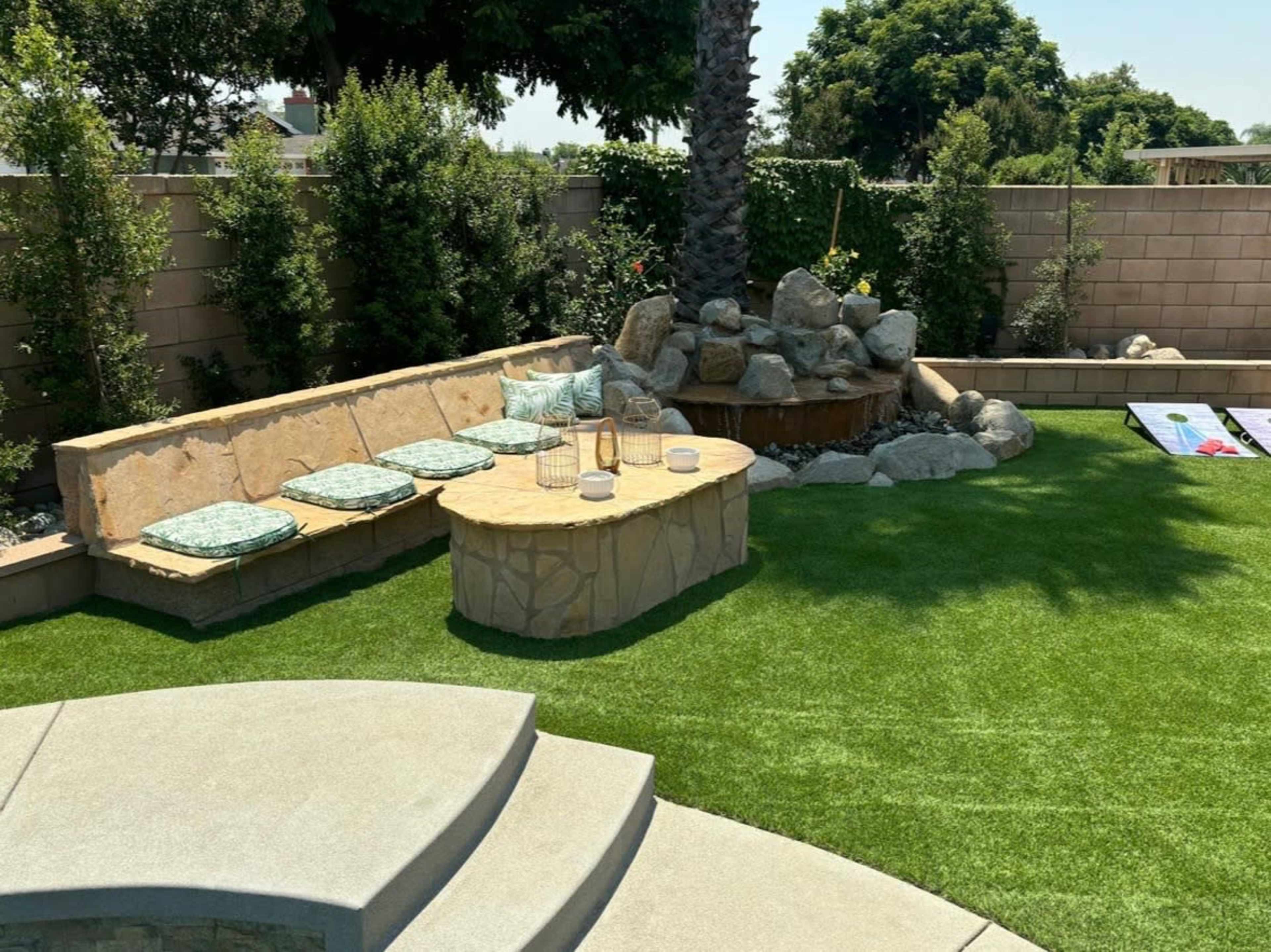 The image shows a landscaped backyard with an artificial lawn, a stone seating area, a round table, and a rock fountain surrounded by greenery.