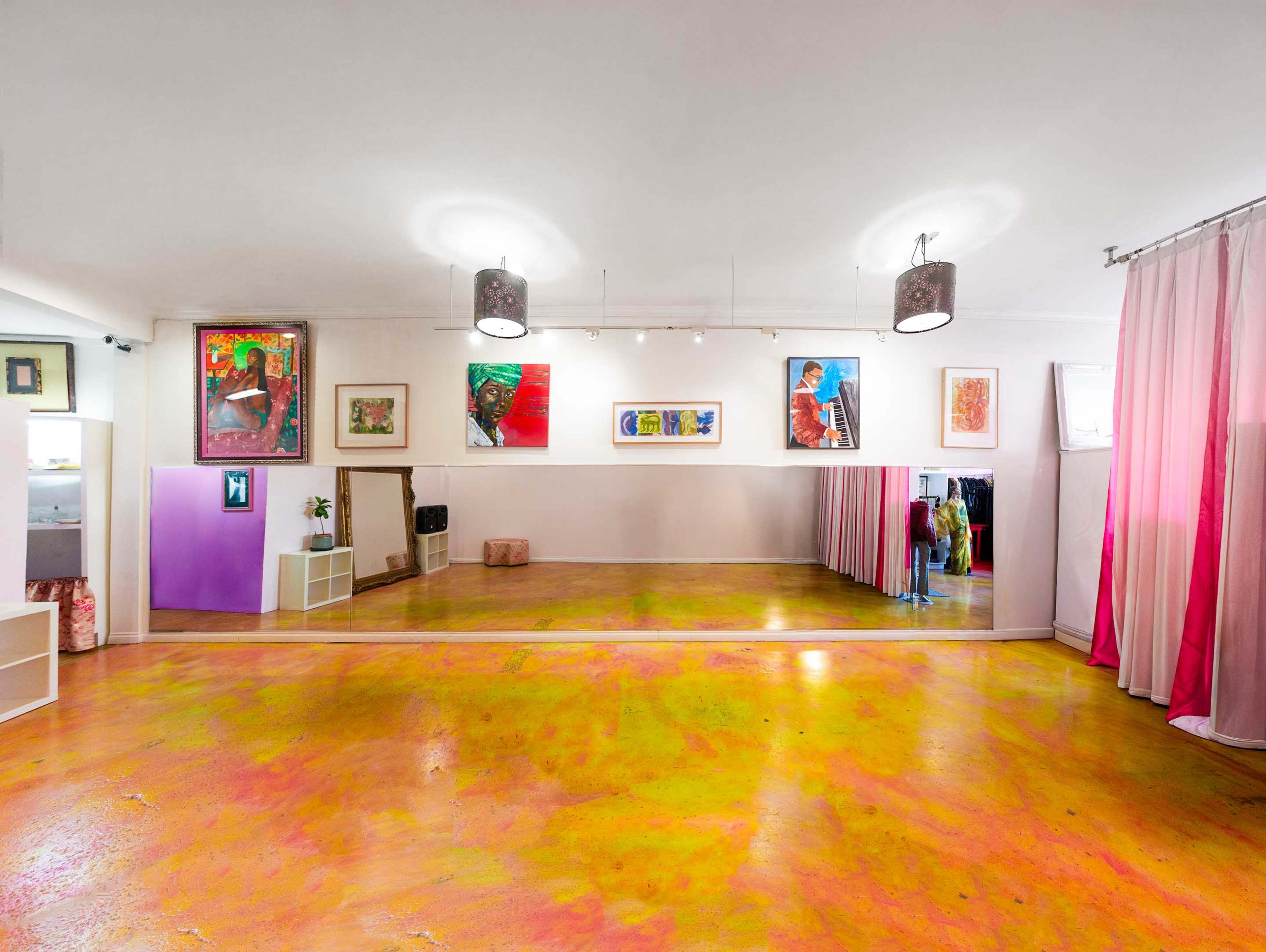 The image shows a spacious art studio with colorful paintings on the walls, a large mirror, and a vibrant, painted floor.