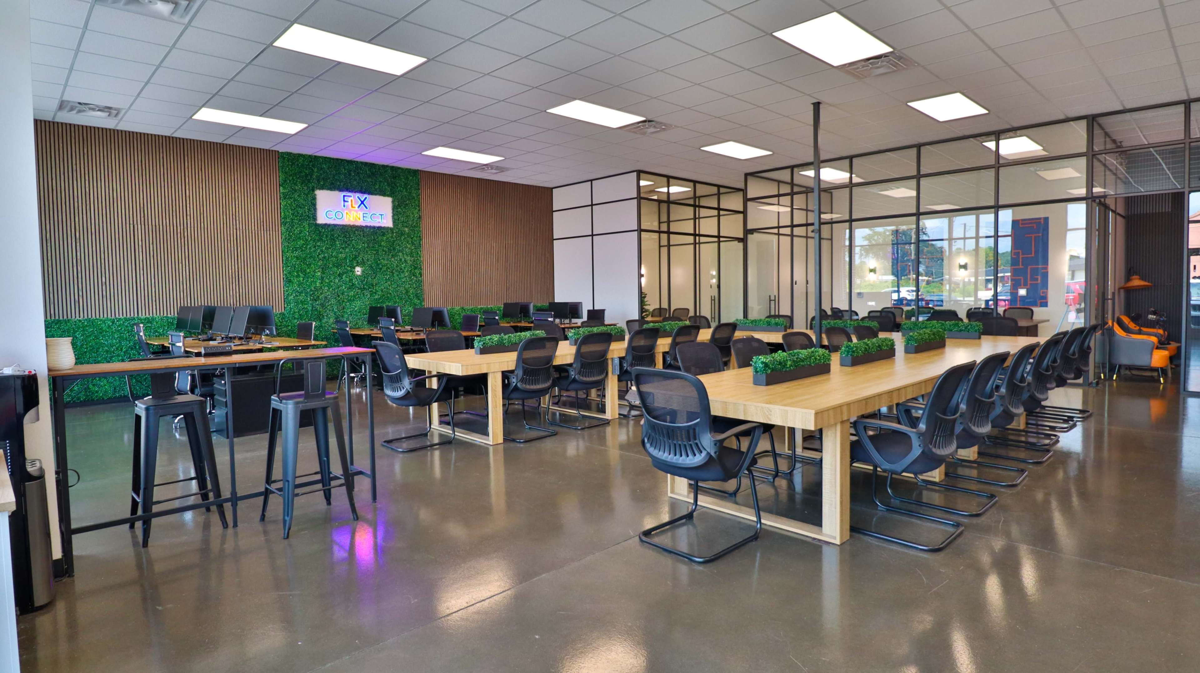 The image shows a spacious coworking area with large tables, black office chairs, and a green wall in the background featuring a sign that reads "FX Coworking."