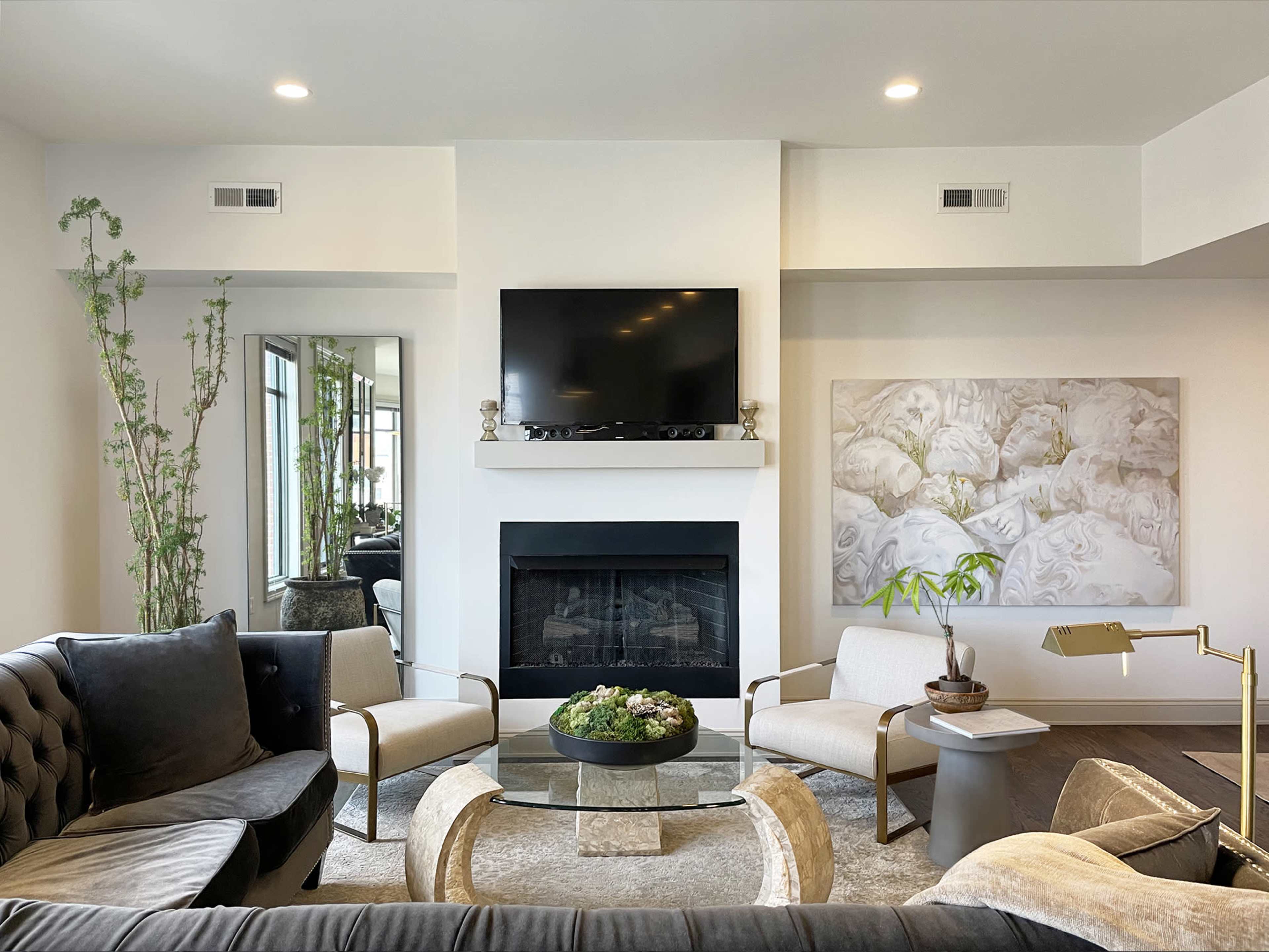 The image shows a modern living room with a fireplace, a large flat-screen TV mounted above it, and a circular glass coffee table surrounded by plush seating and decorative elements.