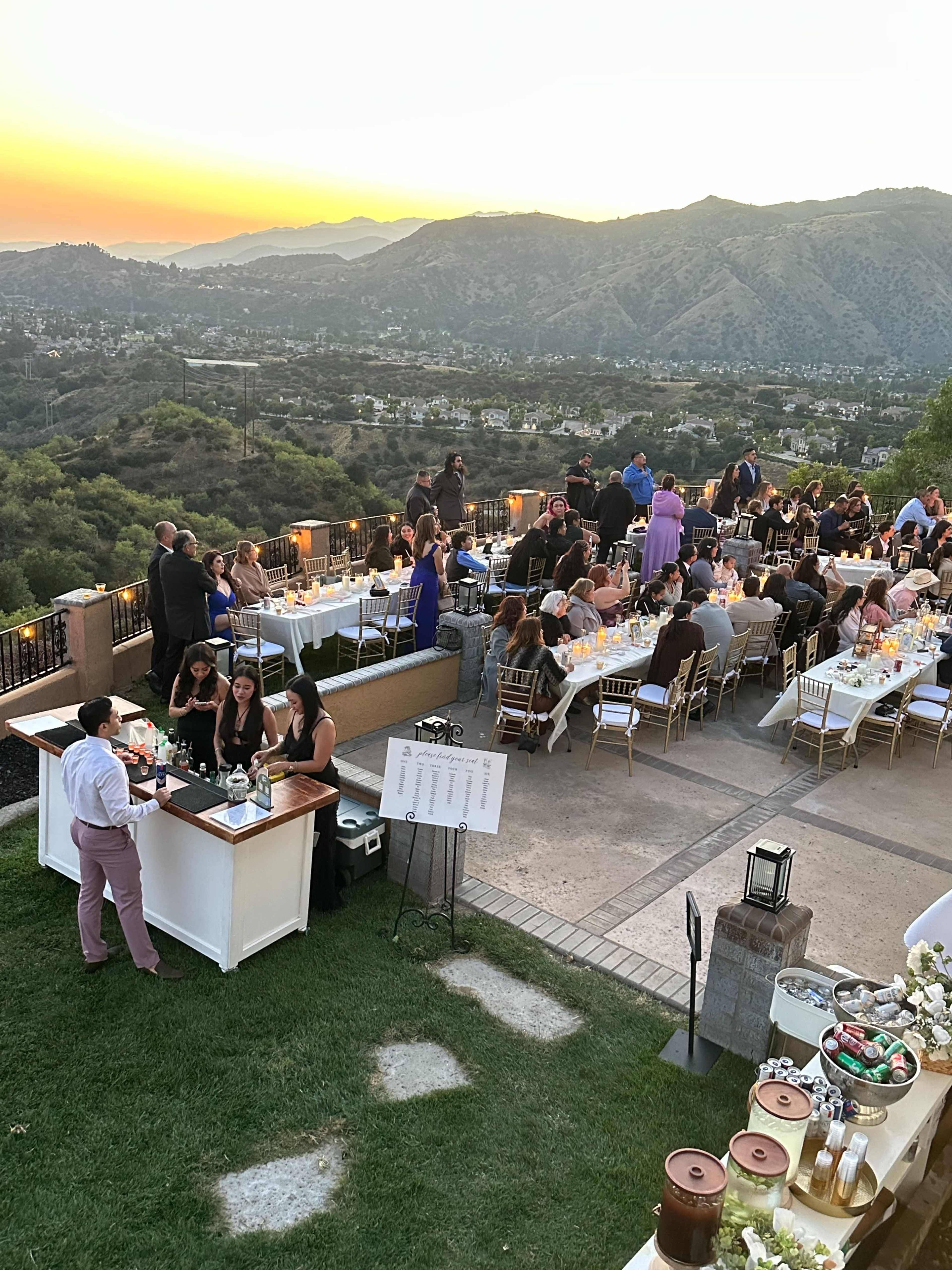 A large outdoor event is taking place at sunset, with guests mingling near elegantly set tables overlooking hills and a valley.