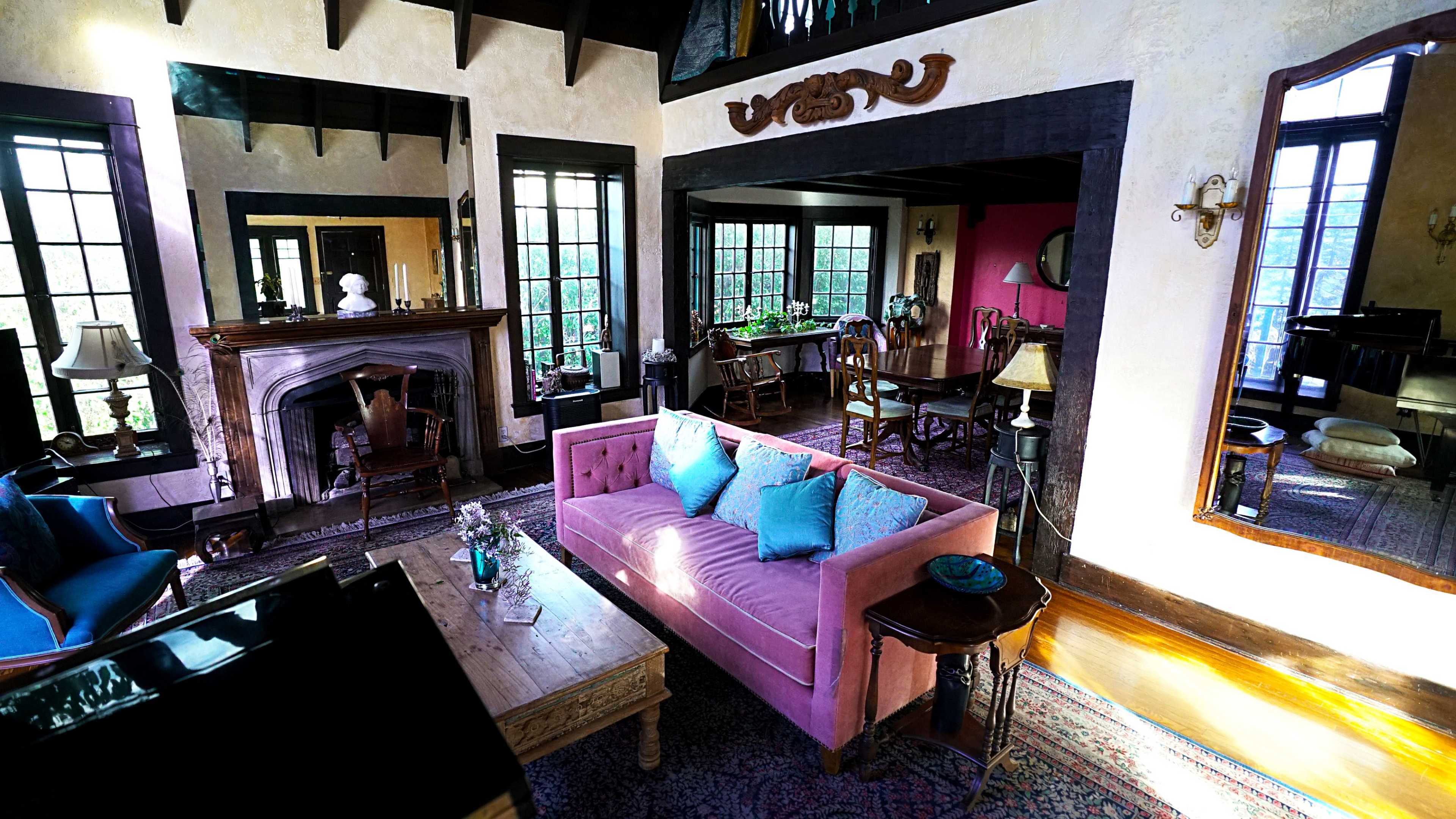 The image shows a spacious living room featuring a pink sofa, a wooden table, a fireplace, and large windows allowing natural light to fill the space.