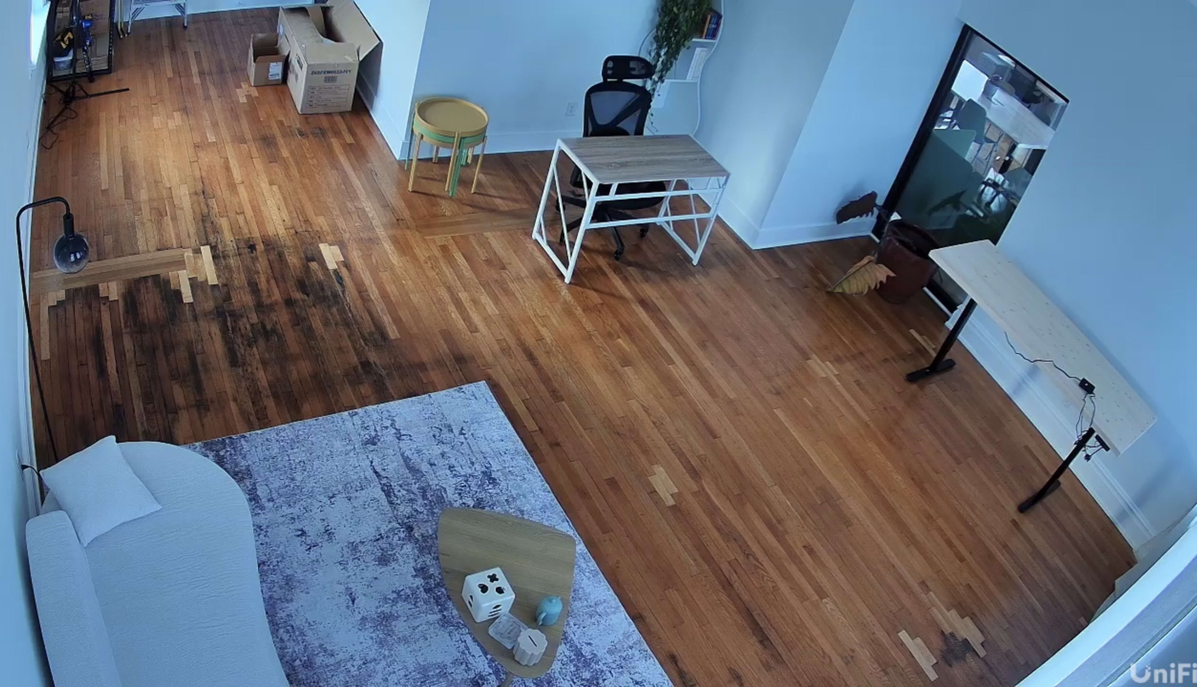 The image shows a partially furnished room with wooden flooring, a desk and chair set, a small stool, and a few boxes in one corner.
