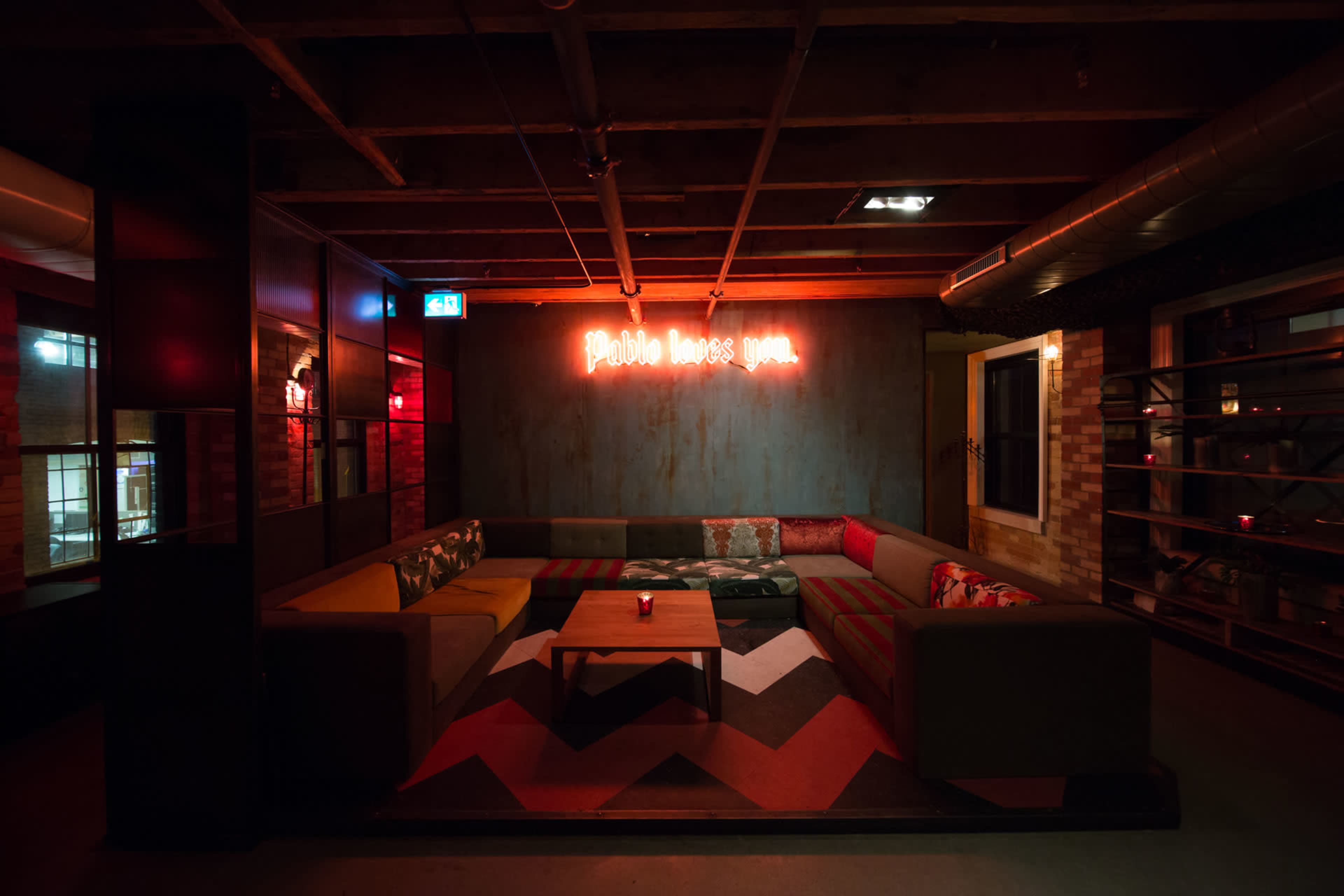 The image shows a modern lounge area with a central sectional sofa, a patterned rug, and a neon sign that reads "Phobe loves you" on the wall.