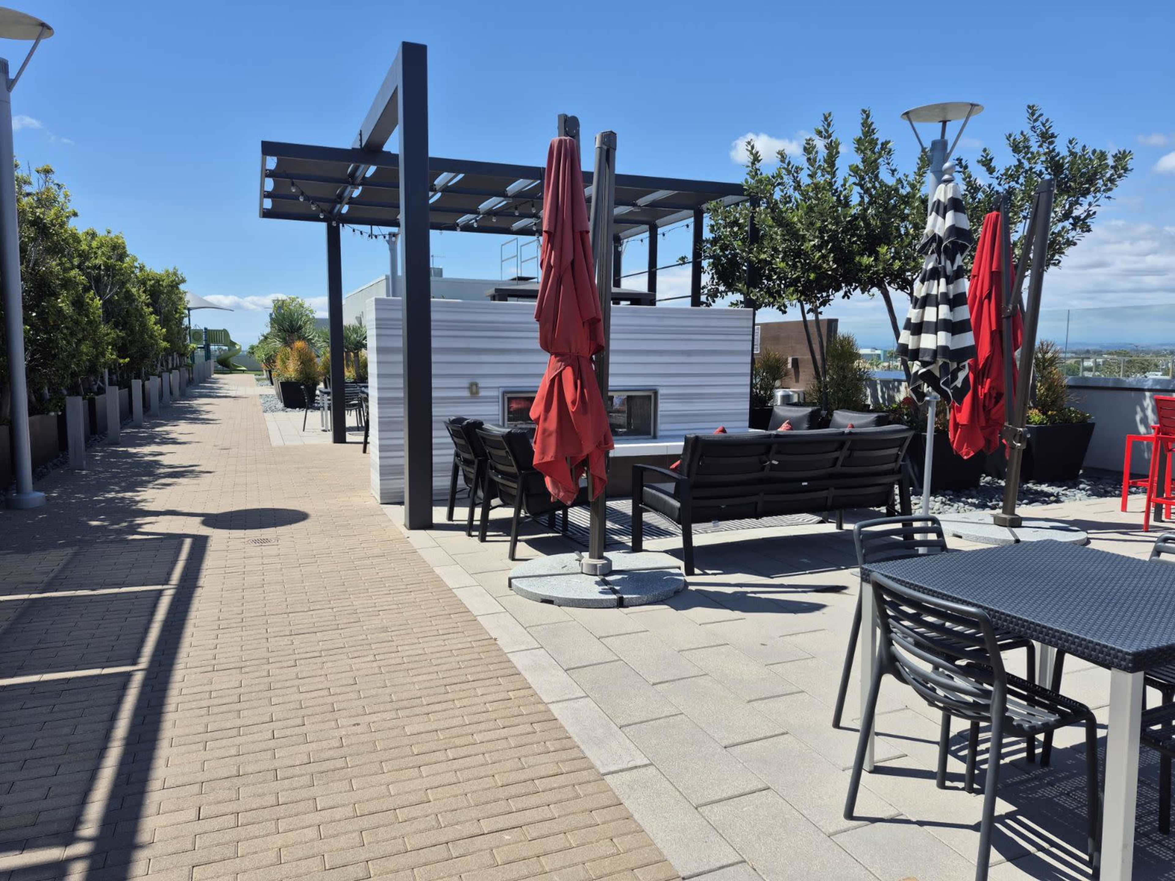 The image shows a rooftop patio featuring wooden seating areas, red umbrellas, and landscaped greenery under a clear blue sky.