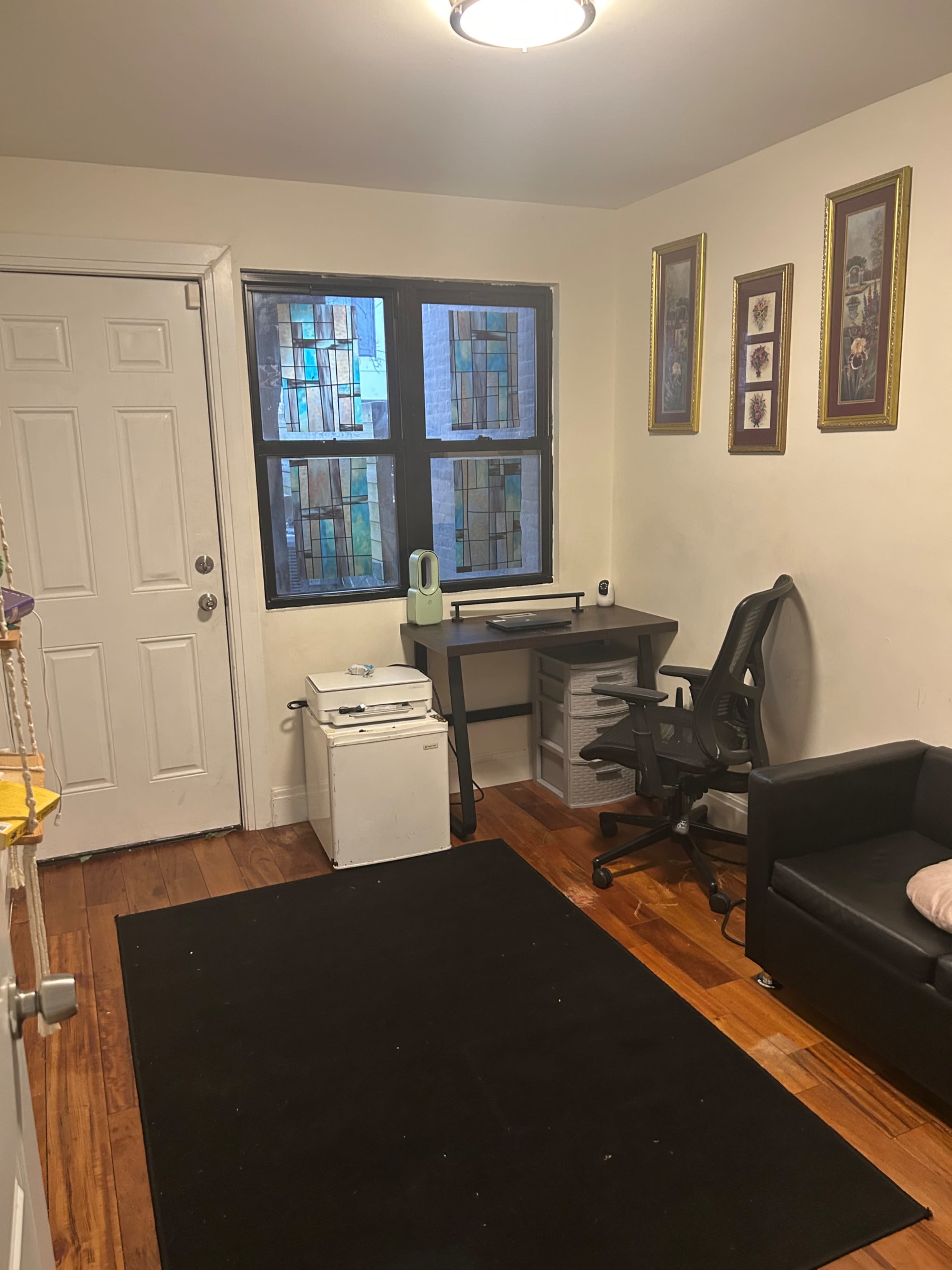 The image shows a small office room containing a desk with a chair, a black rug on the wooden floor, a mini-fridge, and a couch along with two framed pictures on the wall.