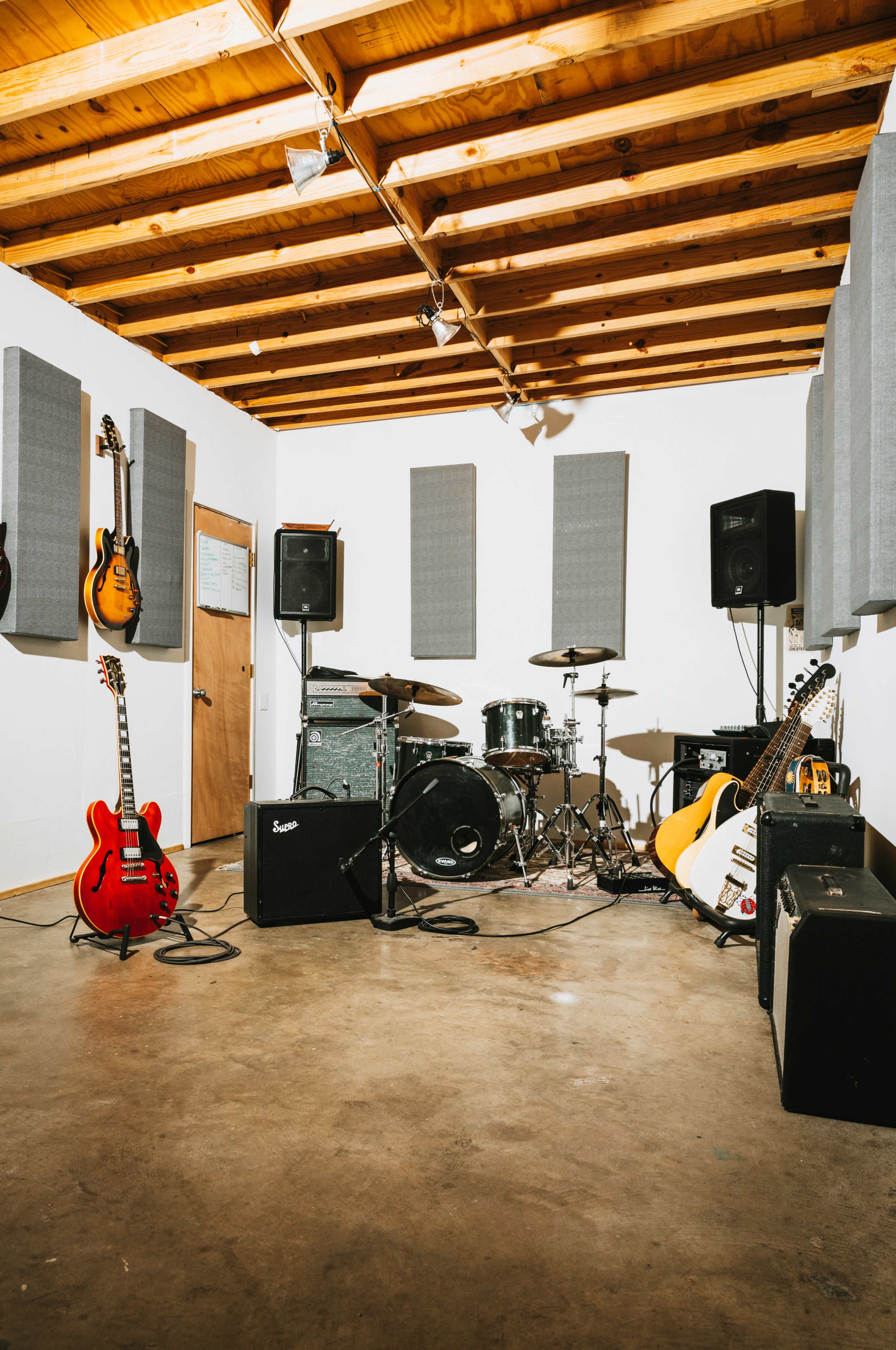 A music rehearsal space featuring guitars on the walls, a drum set, amplifiers, and soundproofing panels.
