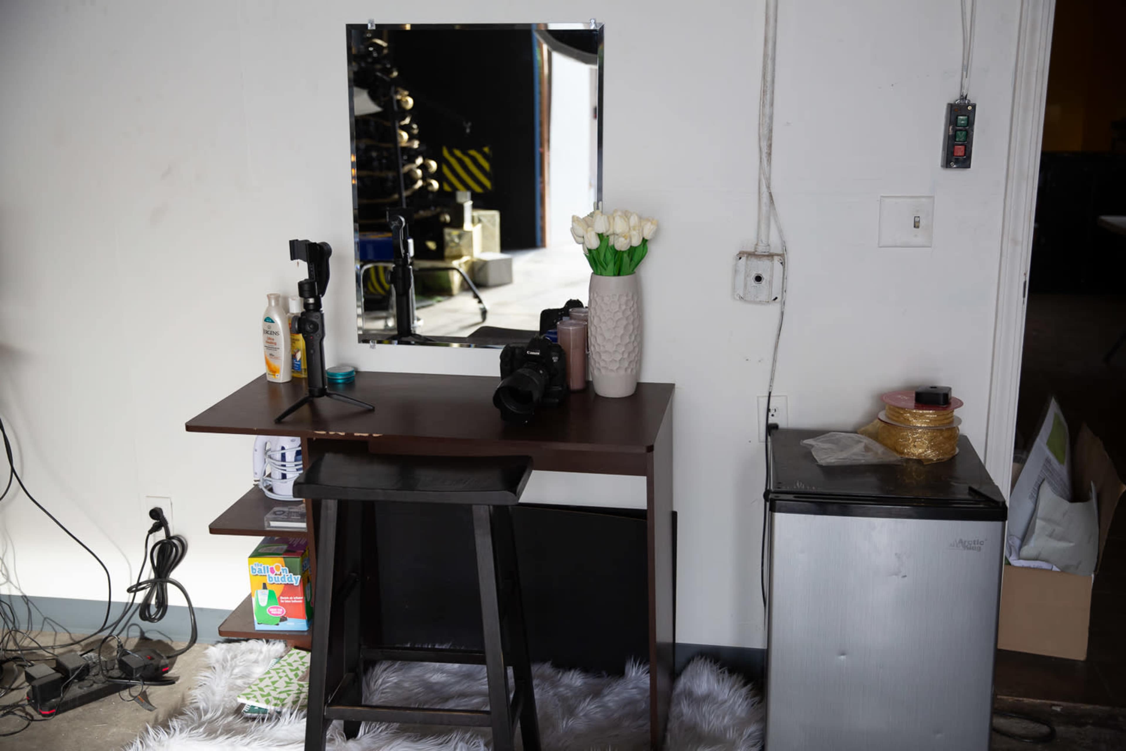 A small wooden vanity with a mirror and a vase of white flowers is positioned against a wall, next to a mini refrigerator and a stack of boxes.