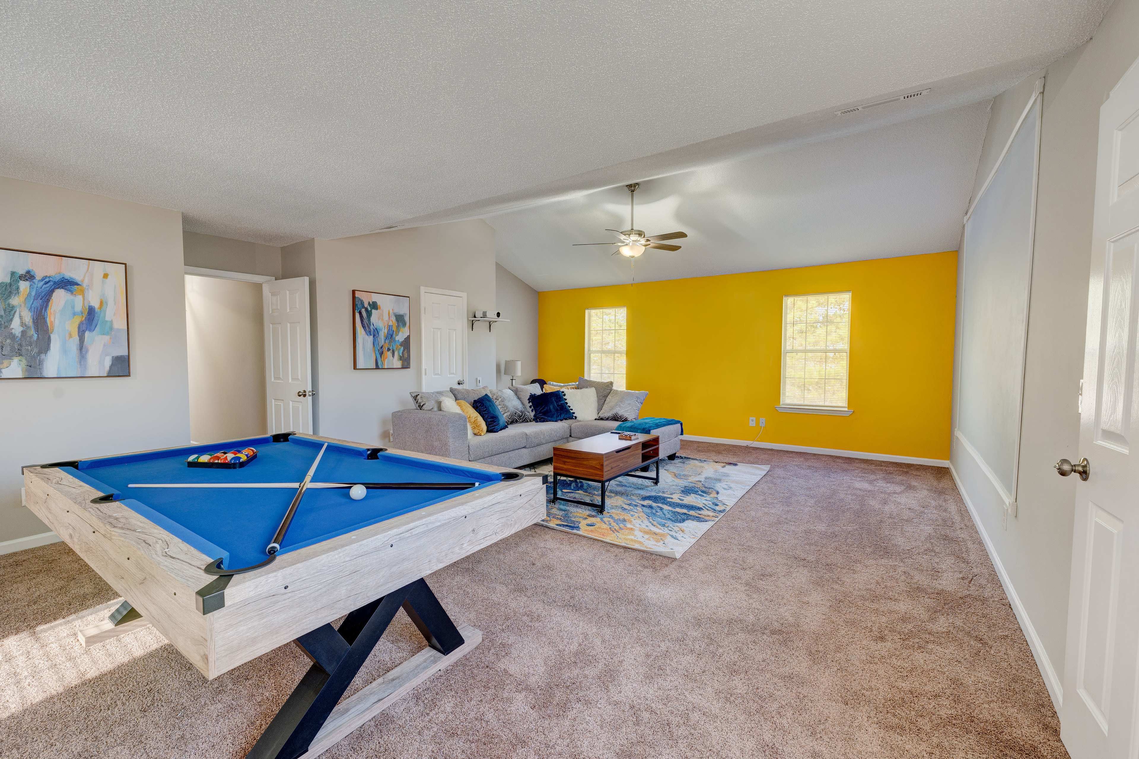 A game room features a blue pool table, a gray sofa, and yellow walls with natural light streaming through the windows.