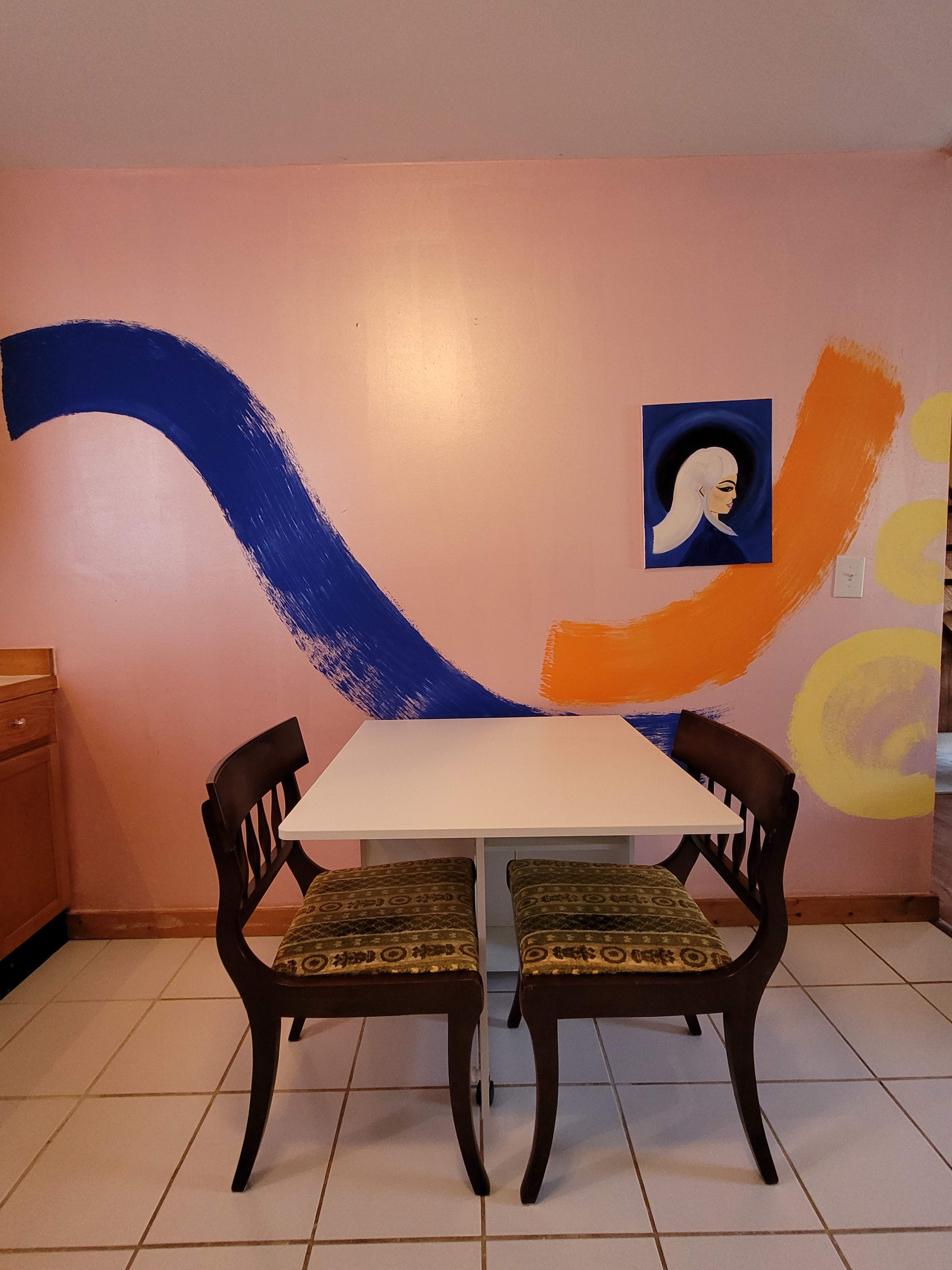 A white table with two dark wooden chairs is positioned in a kitchen with a pink wall featuring abstract painted designs in blue and orange, along with a portrait.