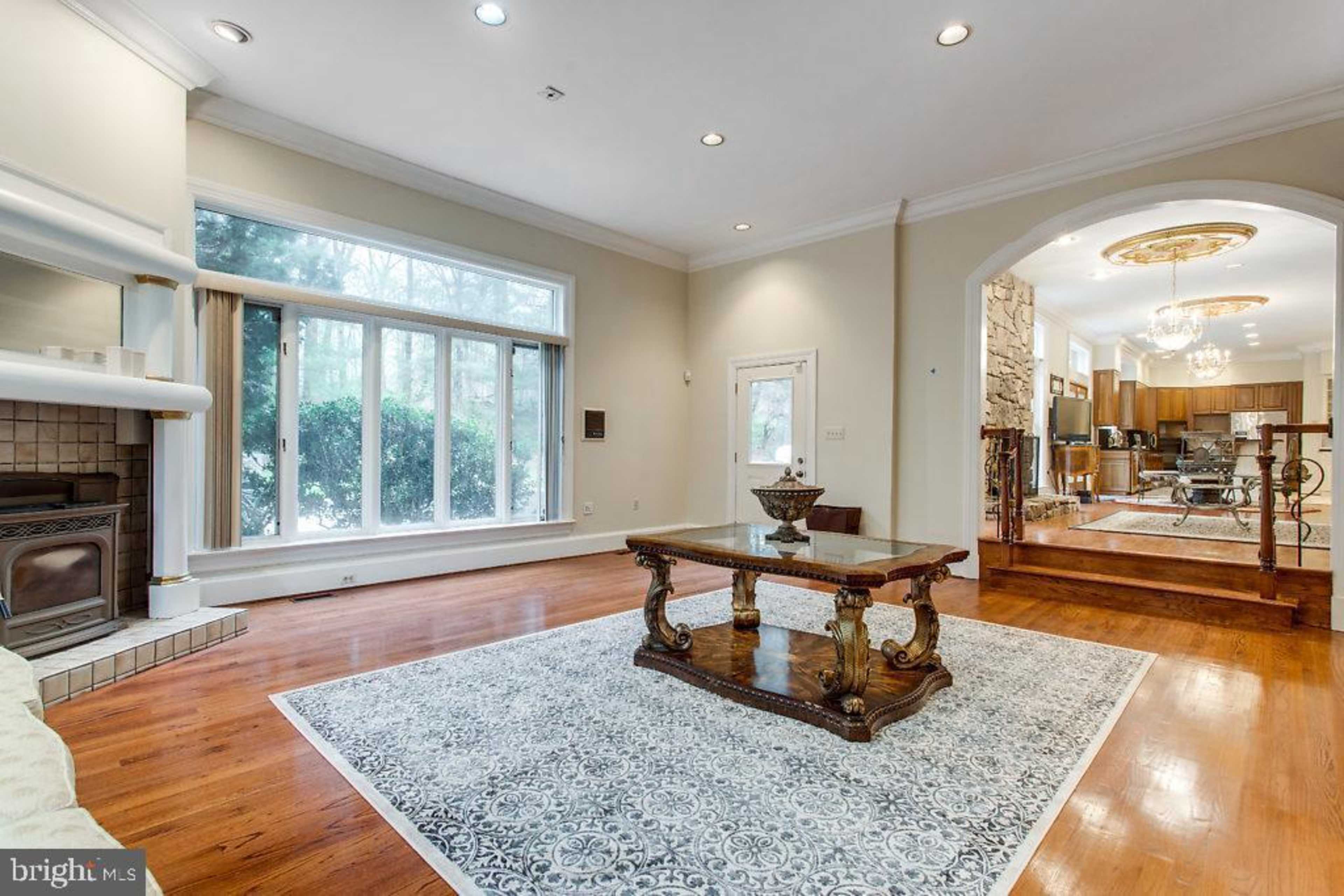 A spacious living room featuring a wooden table on a patterned rug, large windows allowing natural light, and a view of greenery outside.