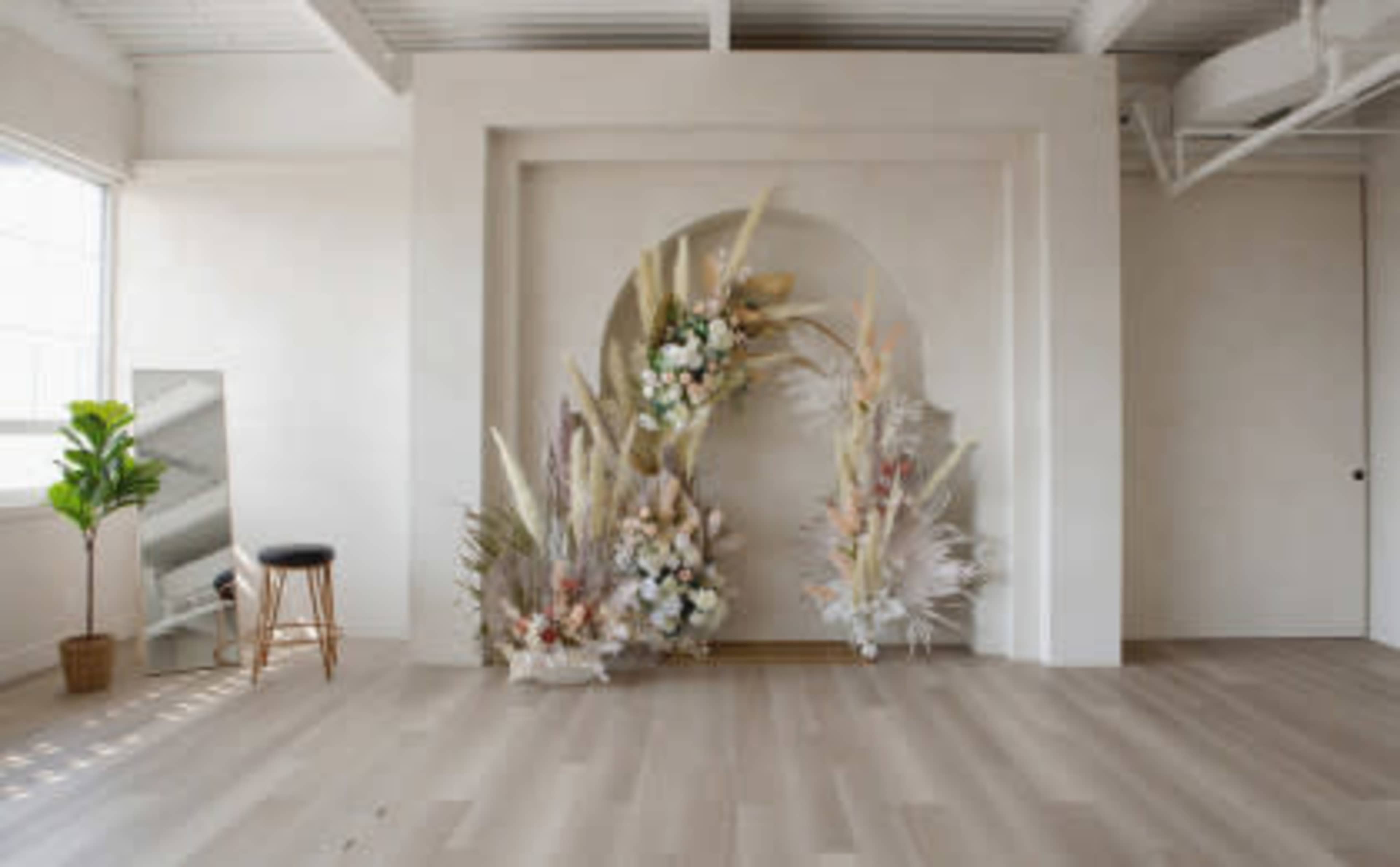 A minimalist interior space with a large arch of decorative dried flowers and plants against a light-colored wall, accompanied by a mirror and a stool.