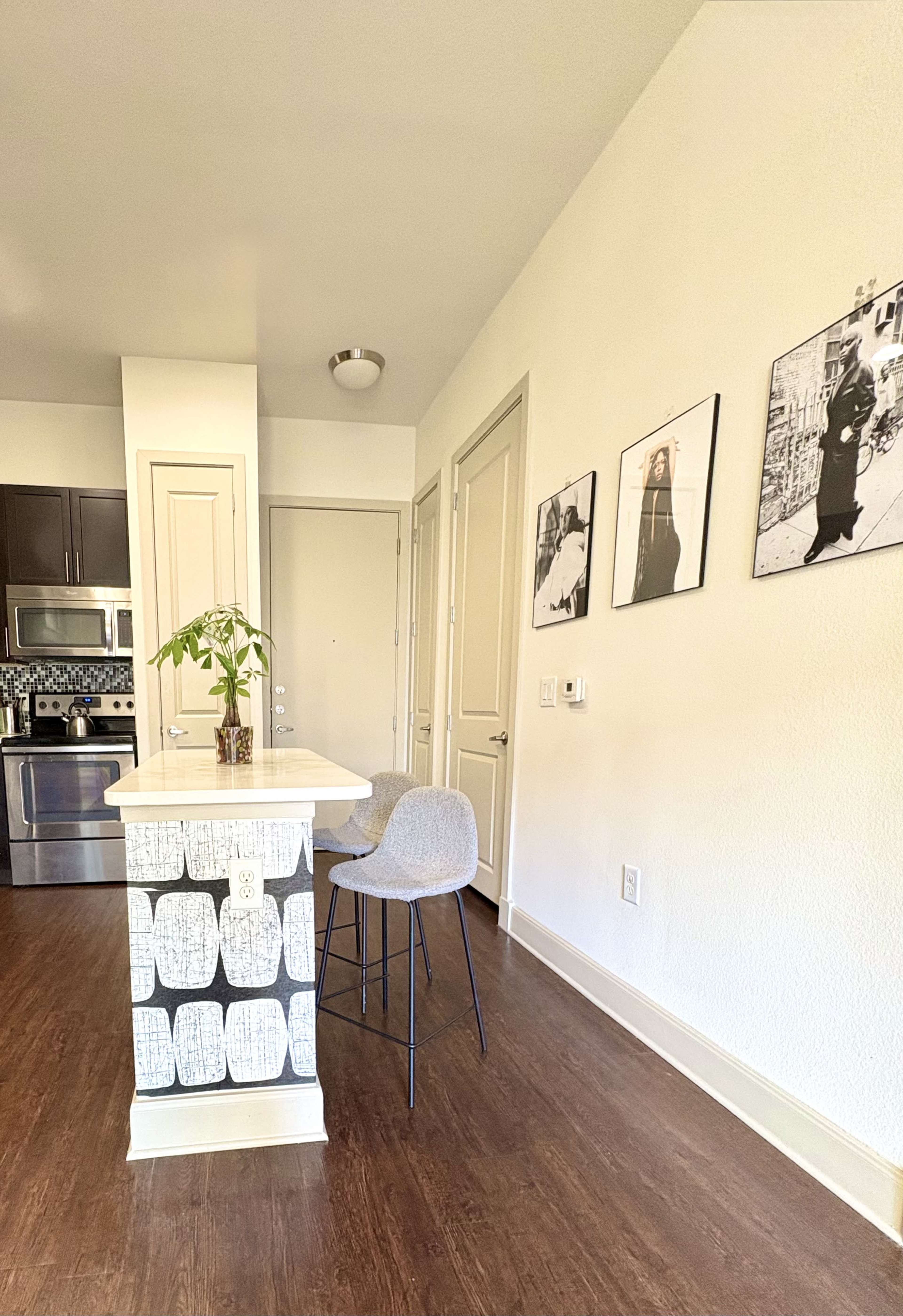 The image shows a modern kitchen area with a small island, two bar stools, and framed black-and-white photographs on the wall.