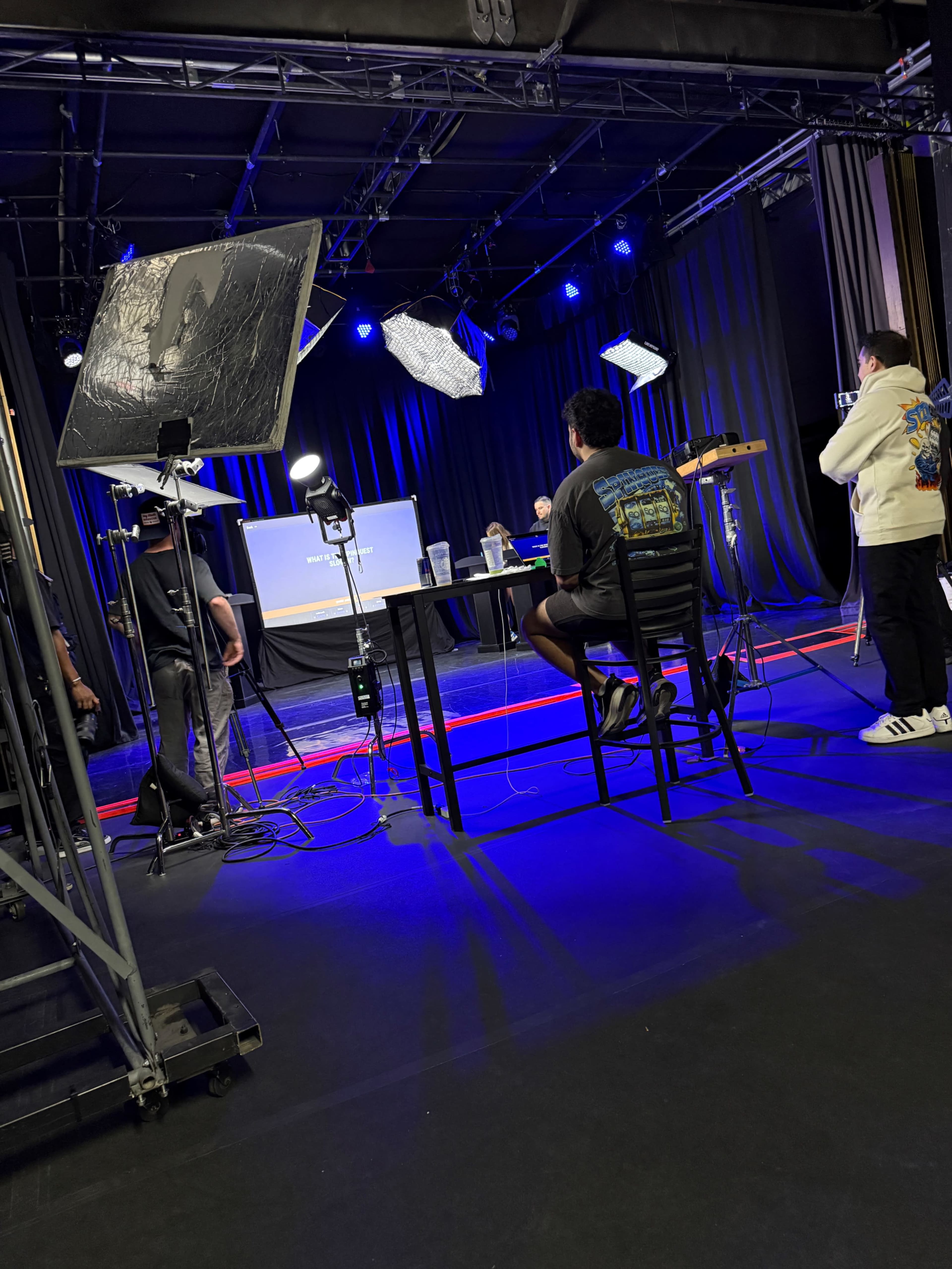 A group of people is set up in a studio with lights, cameras, and a screen displaying information, while one individual sits at a table facing the screen.