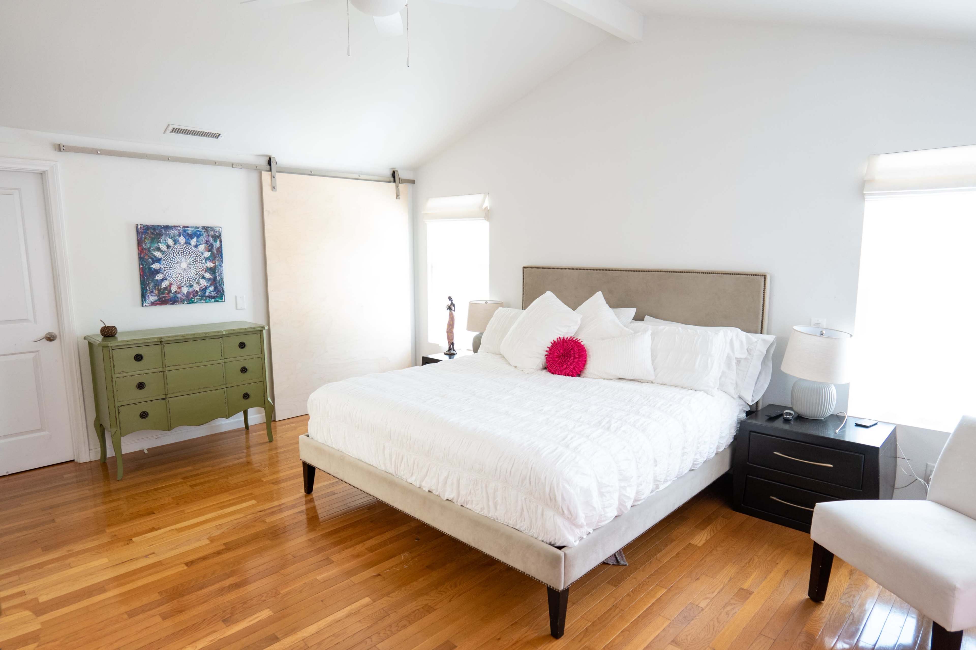 The brightly lit bedroom features a queen-sized bed with white bedding, a green dresser, and large windows.