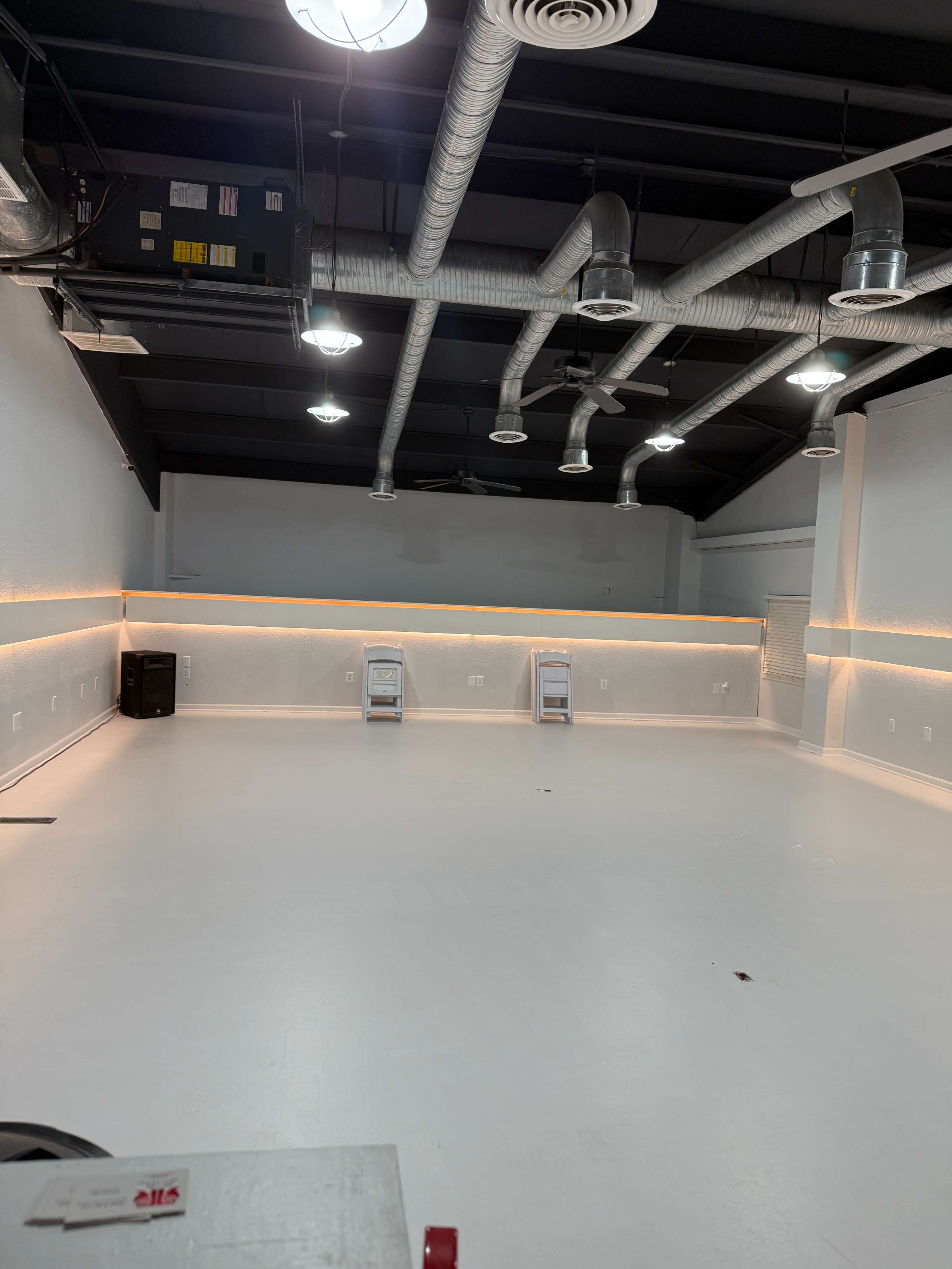 The image shows an empty indoor space with a smooth white floor, exposed ductwork, and minimal lighting along the walls.