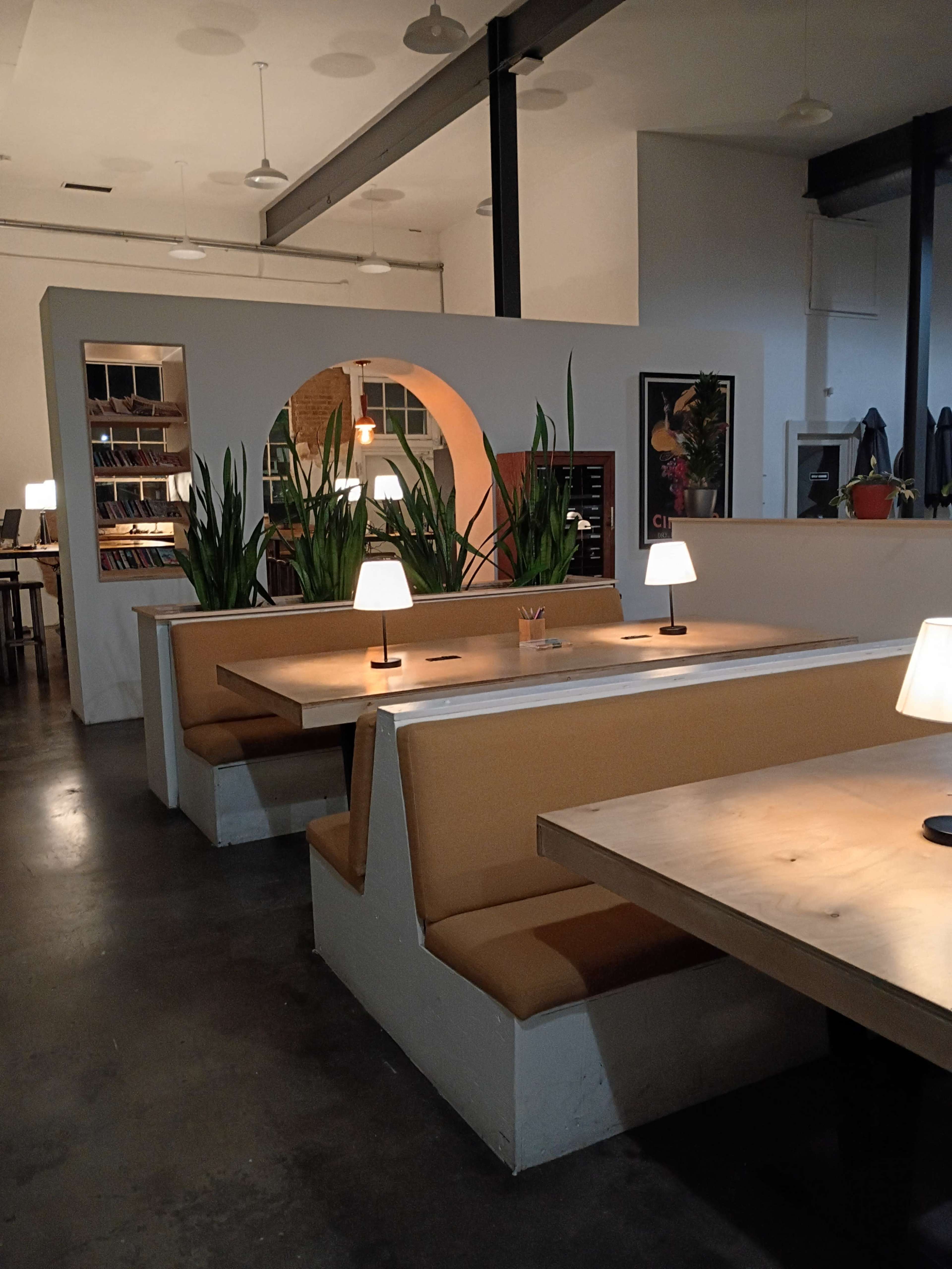 The image shows a modern cafe interior with wooden tables, cushioned benches, and potted plants, illuminated by soft table lamps.