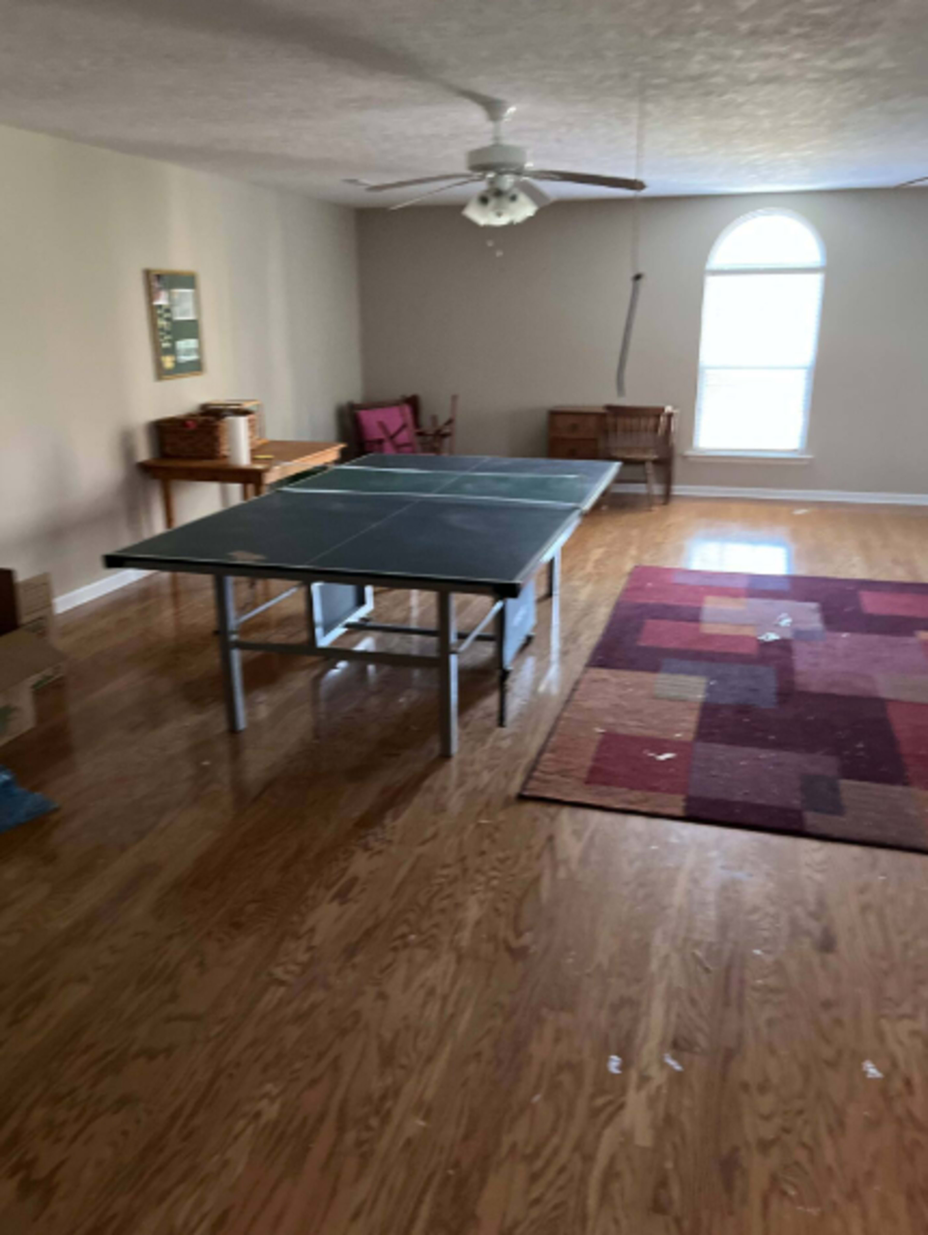 A ping pong table is situated in a sparsely furnished room with wooden floors and a large window.