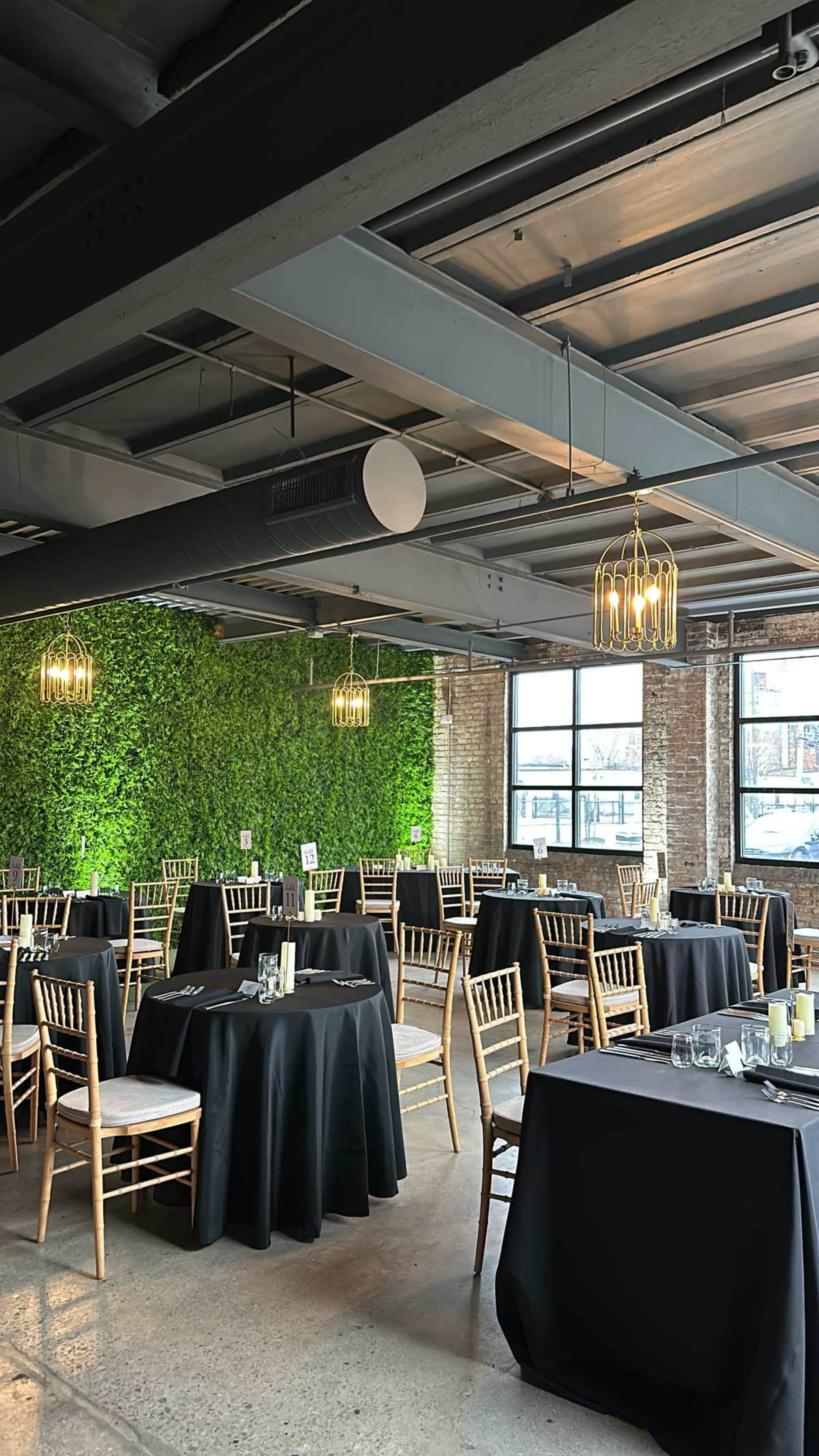 A stylish event space features circular tables with black tablecloths, gold chandeliers, and a green plant wall.