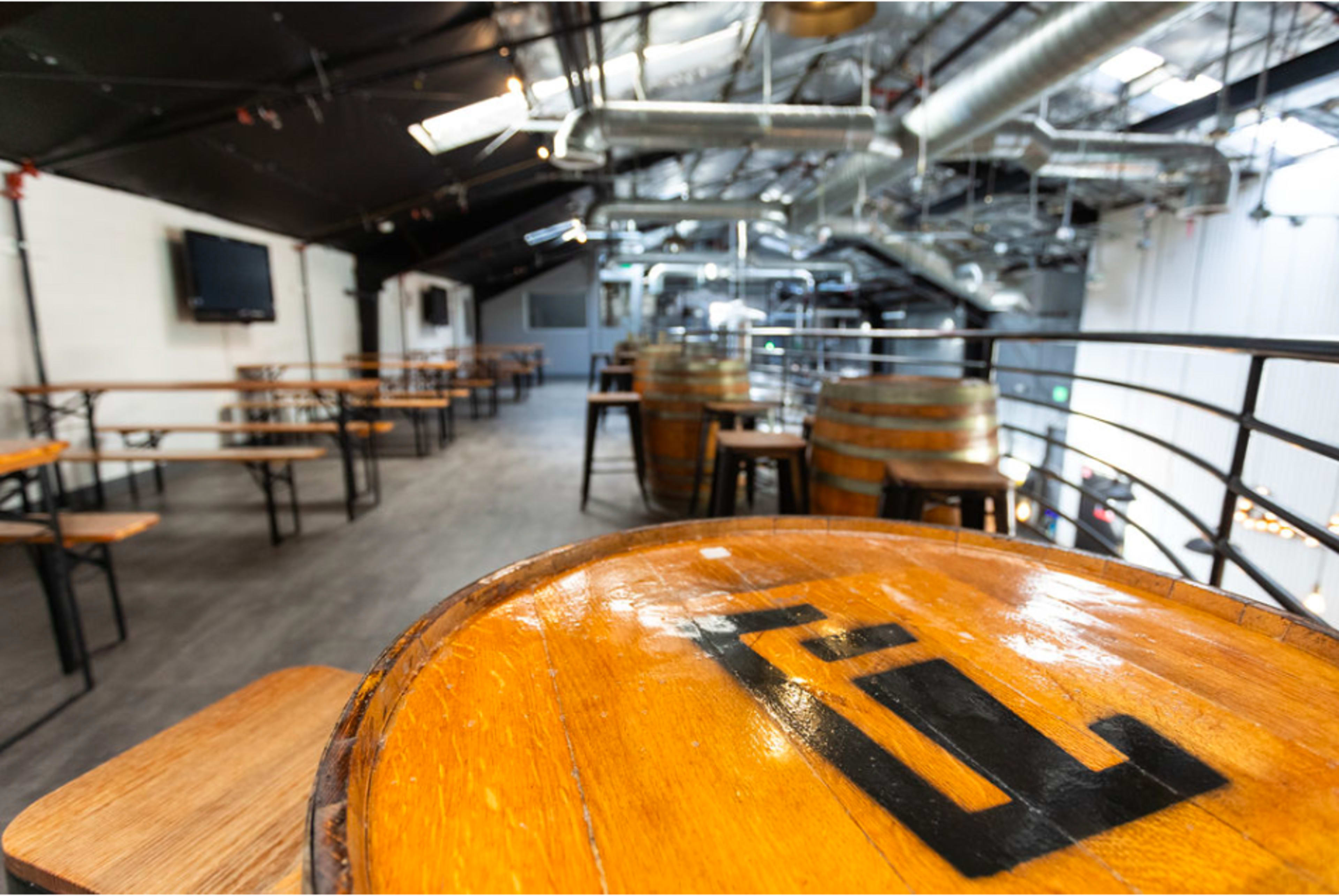 An interior of a bar with wooden tables and barrels, featuring exposed metal ductwork and a TV on the wall.