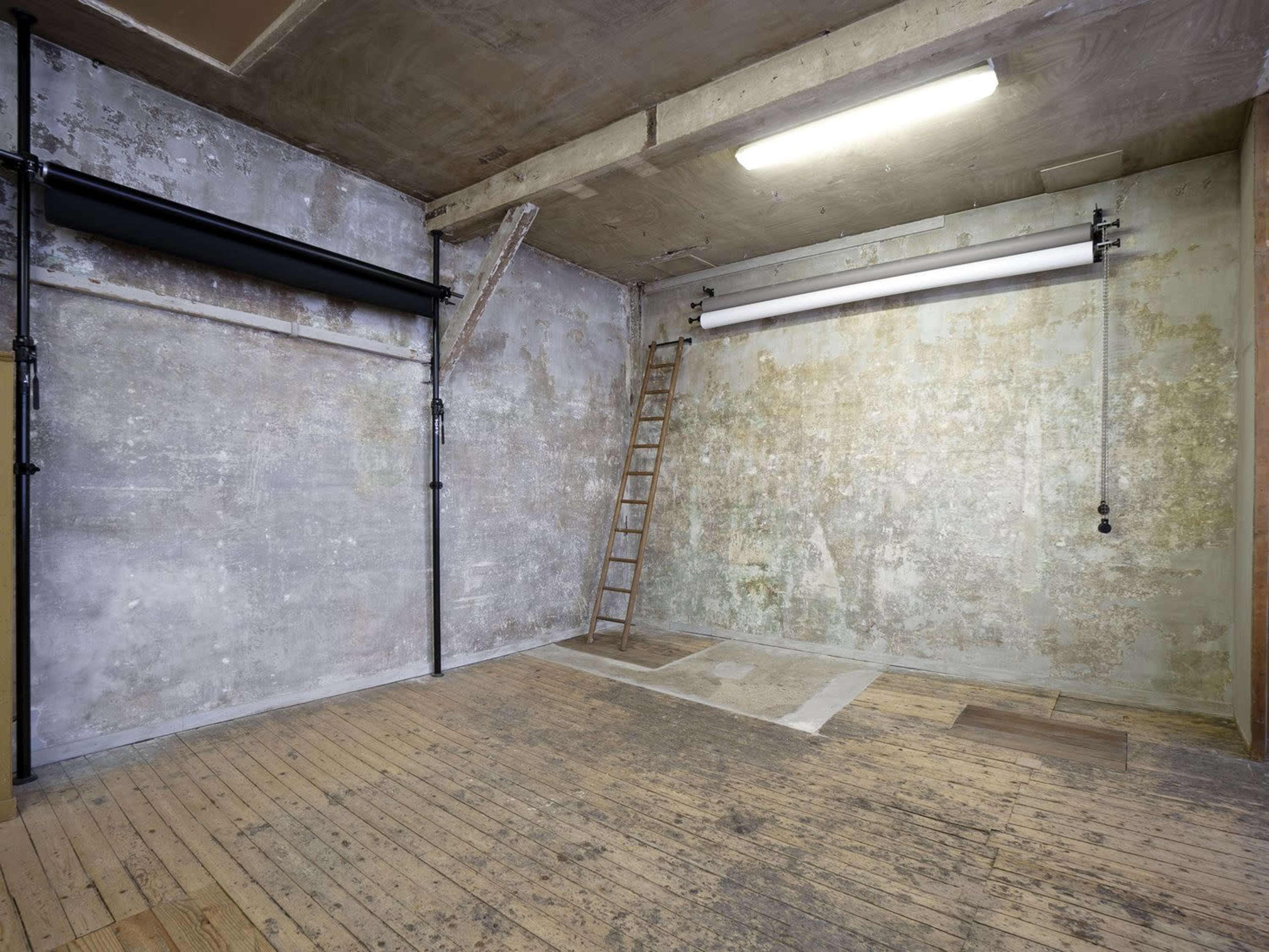 The image shows an empty room with exposed walls, a wooden ladder against one wall, and two ceiling-mounted backdrop supports.