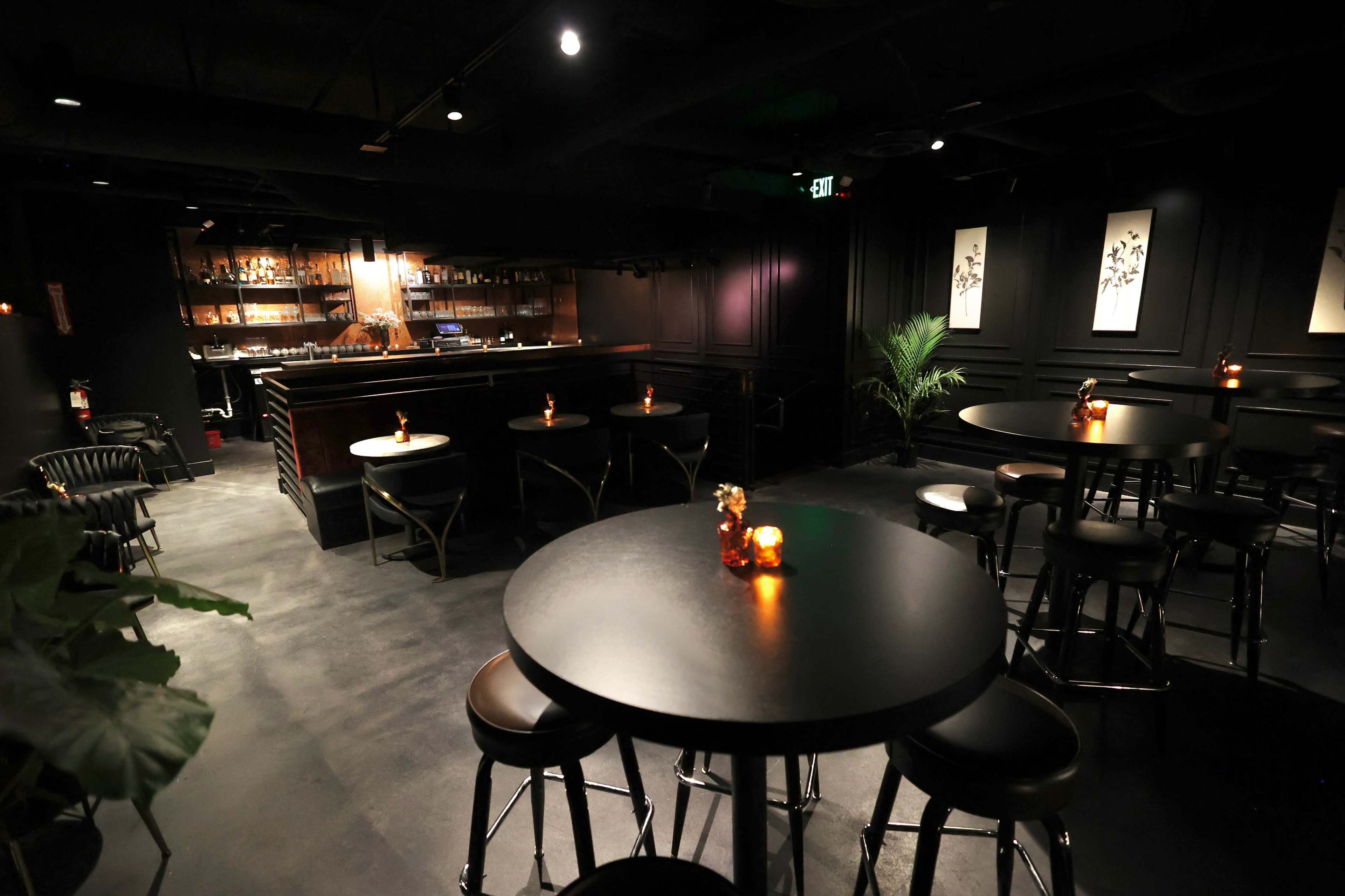 The image shows a dimly lit bar interior featuring several black tables and stools arranged around a central area, with a bar counter in the background and candlelight on the tables.