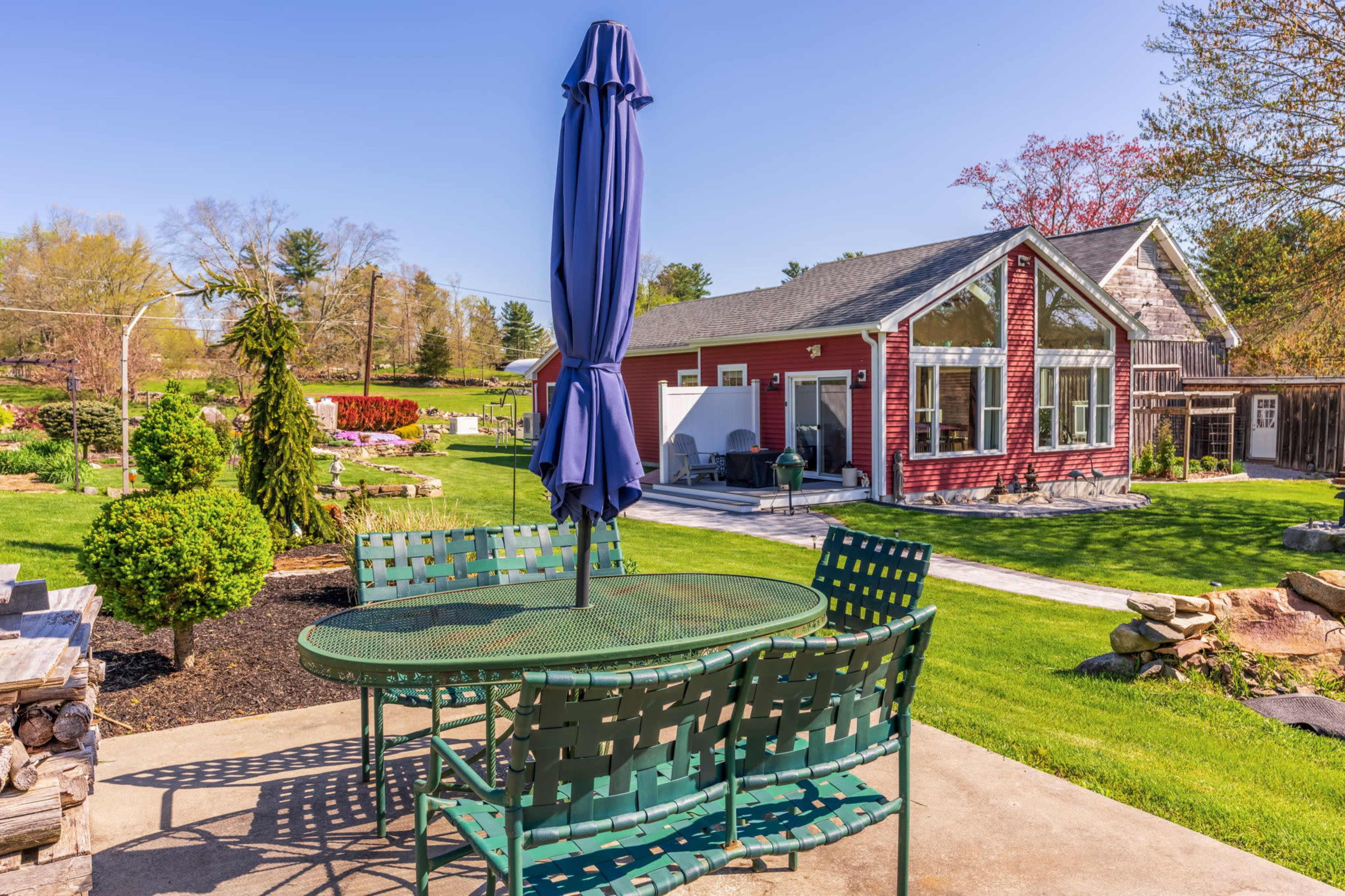 A green metal patio table with chairs and a blue umbrella is set on a concrete surface in a yard featuring a red house and landscaped gardens.