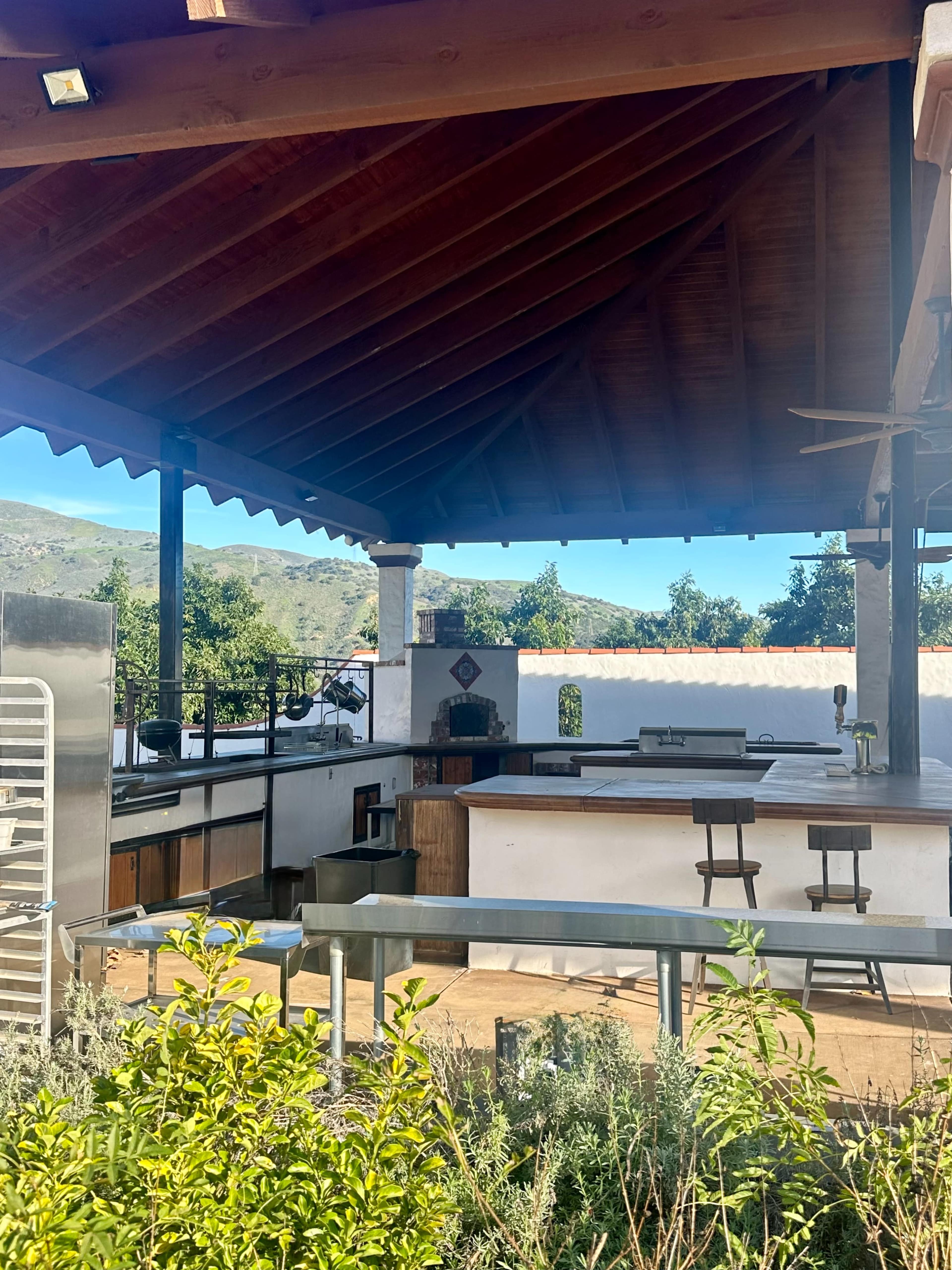 The image shows an outdoor kitchen area with a wooden roof, countertop space, and a few bar stools, surrounded by greenery and mountains in the background.