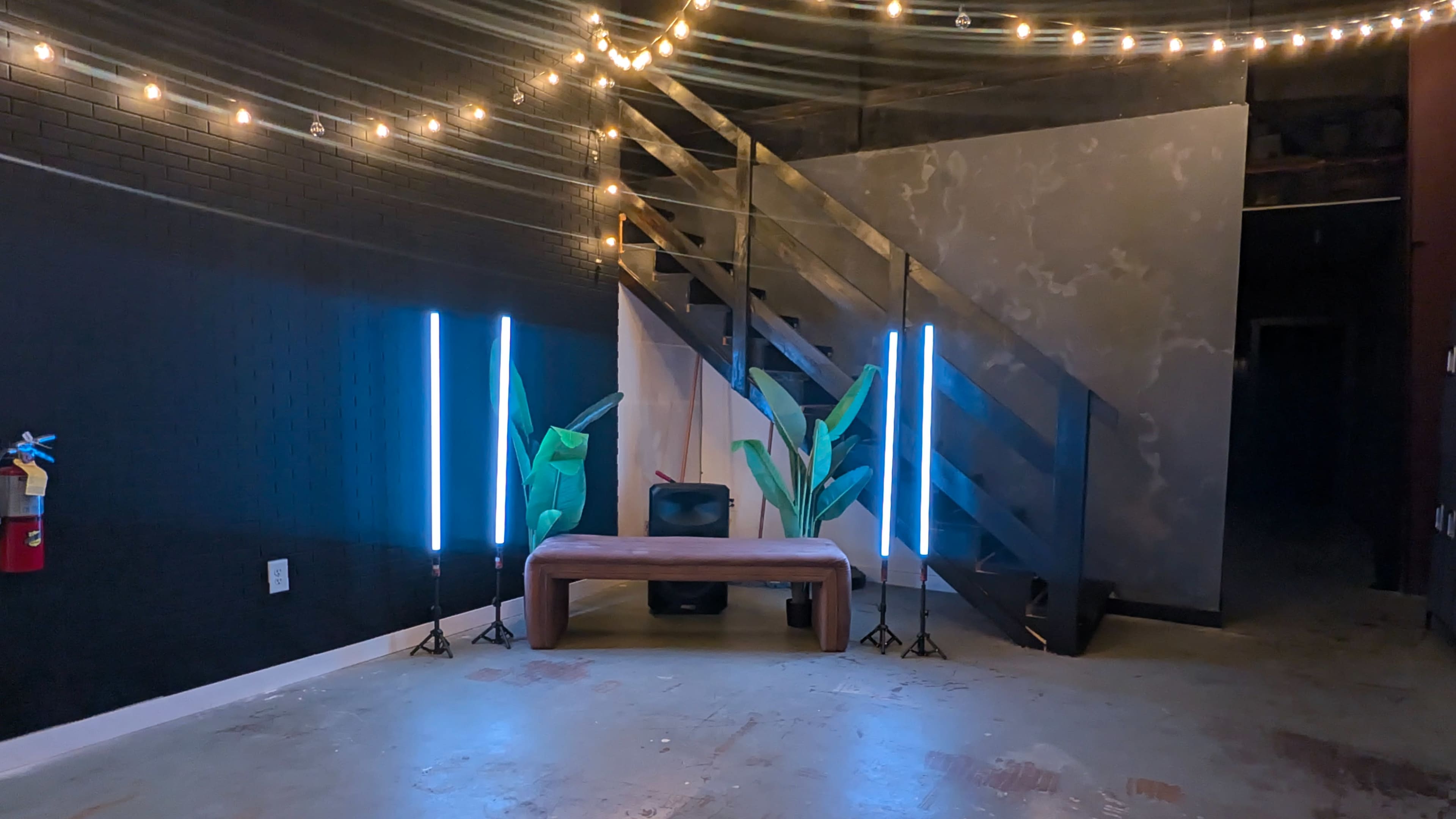 The image shows a dimly lit room with a wooden bench, two illuminated vertical lights, and decorative plants near a staircase.