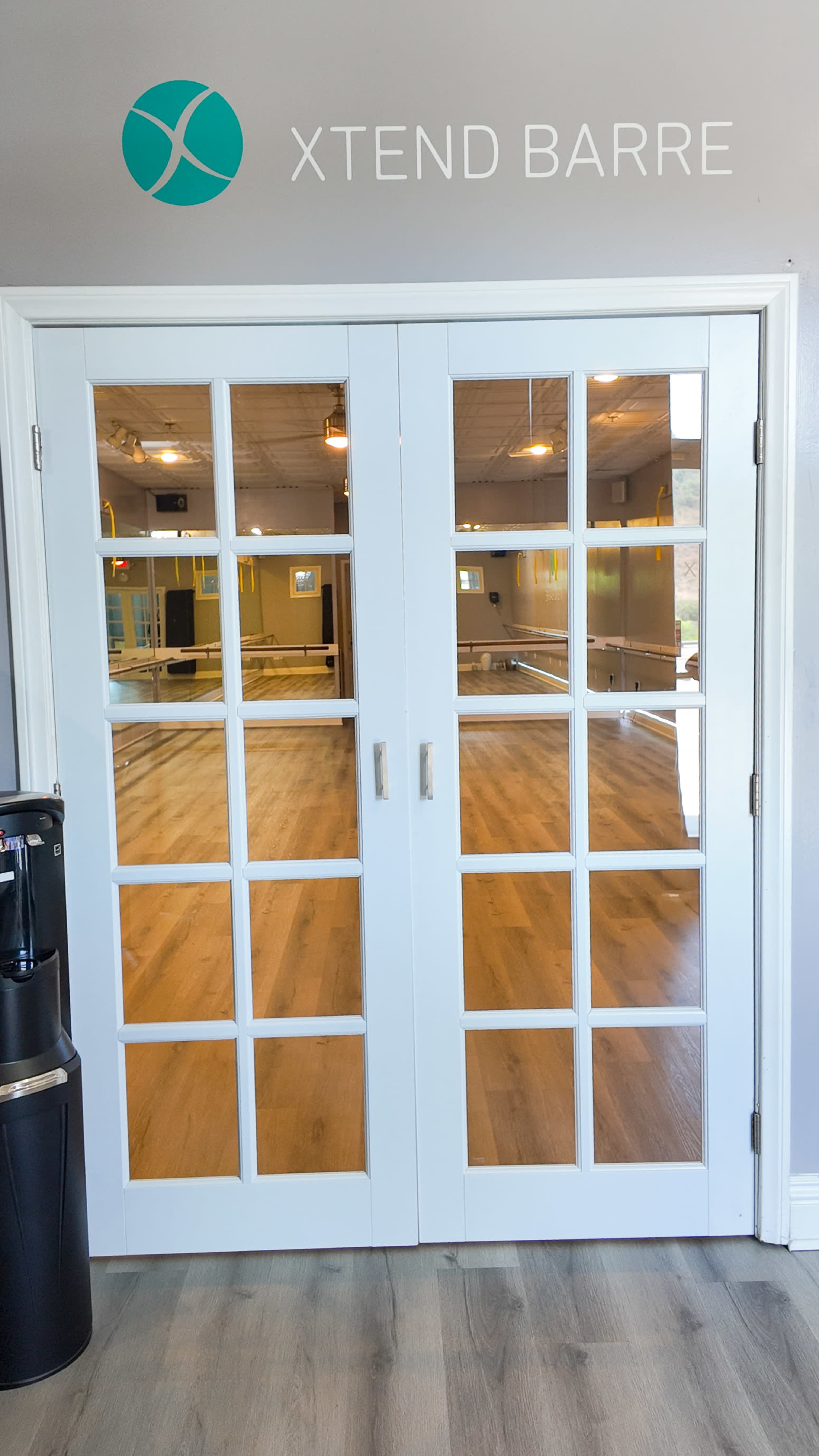 The image shows a set of double doors with glass panes leading to a spacious exercise studio, featuring wooden floors and mirrors.