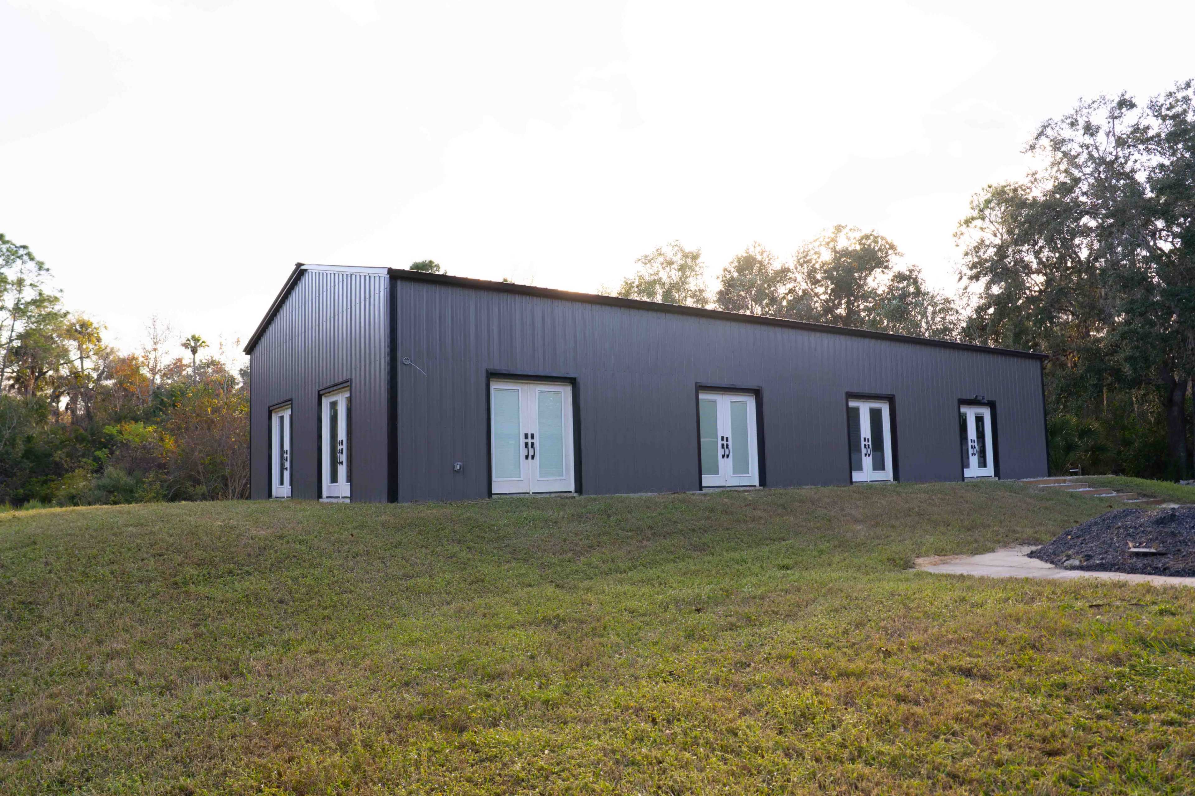 A gray metal building with multiple white doors stands on a grassy lot surrounded by trees.