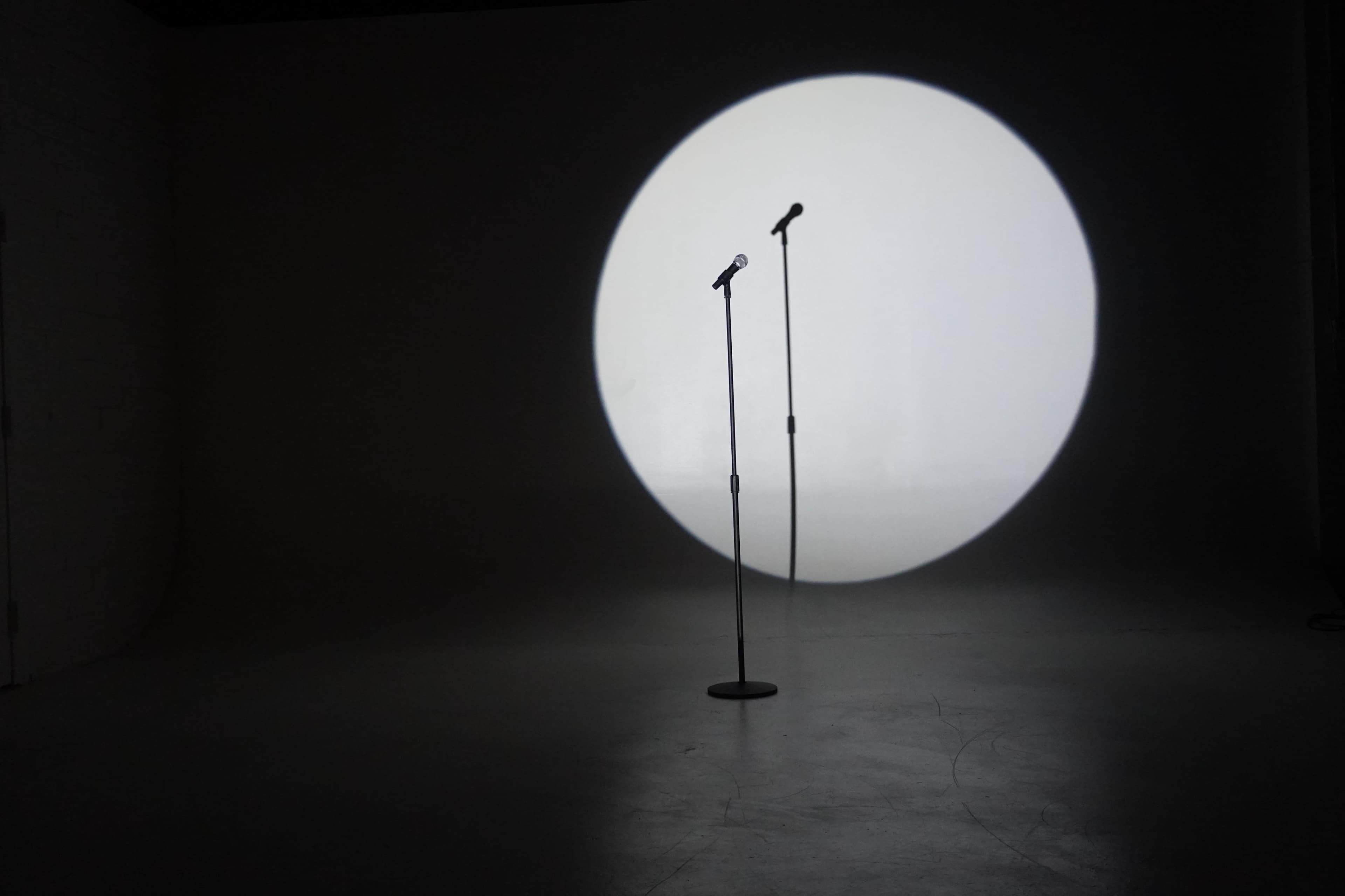 Two microphones are positioned on stands in the center of a spotlight against a plain wall.