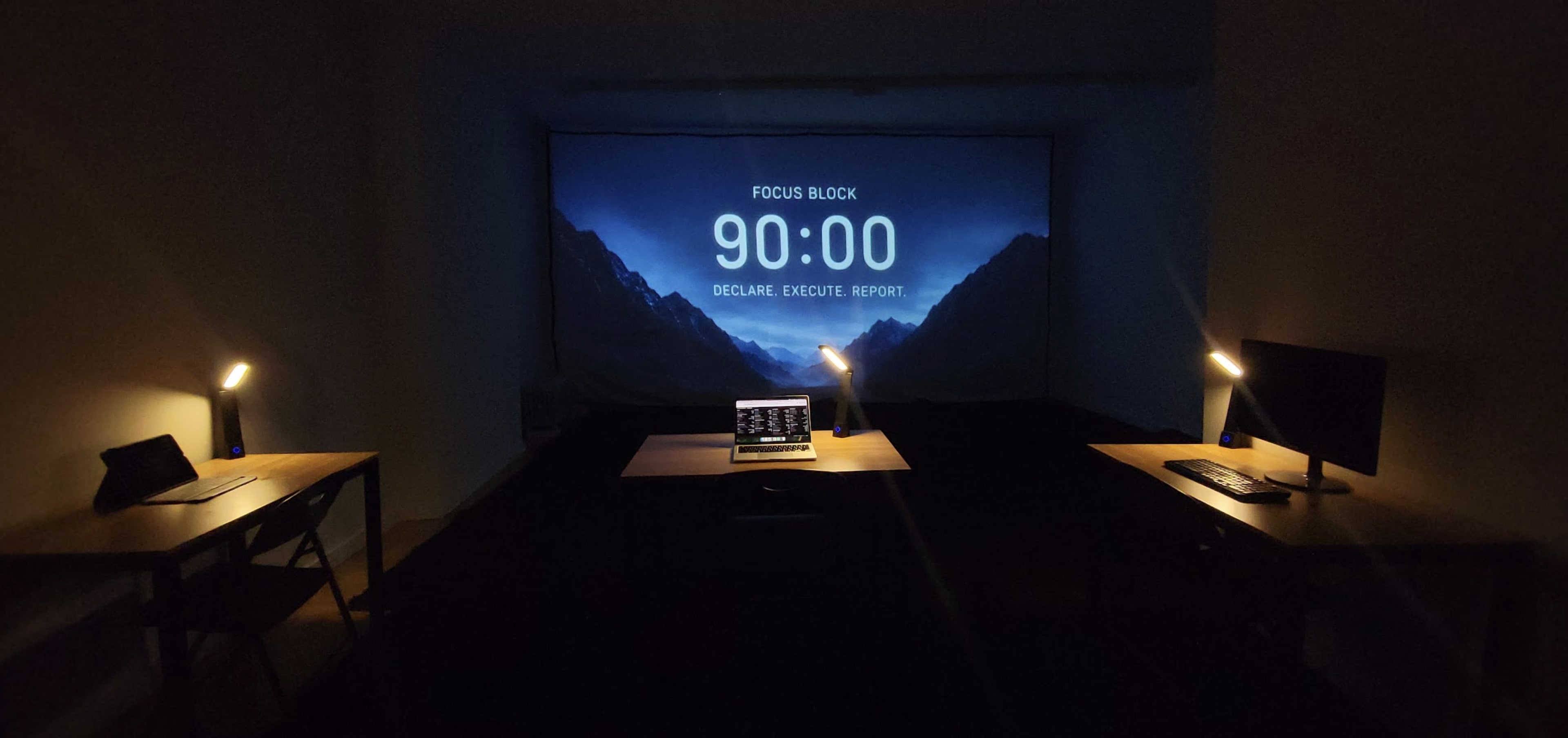 A dimly lit room features two desks with lamps and computers facing a large projection screen displaying a timer set to 90:00.