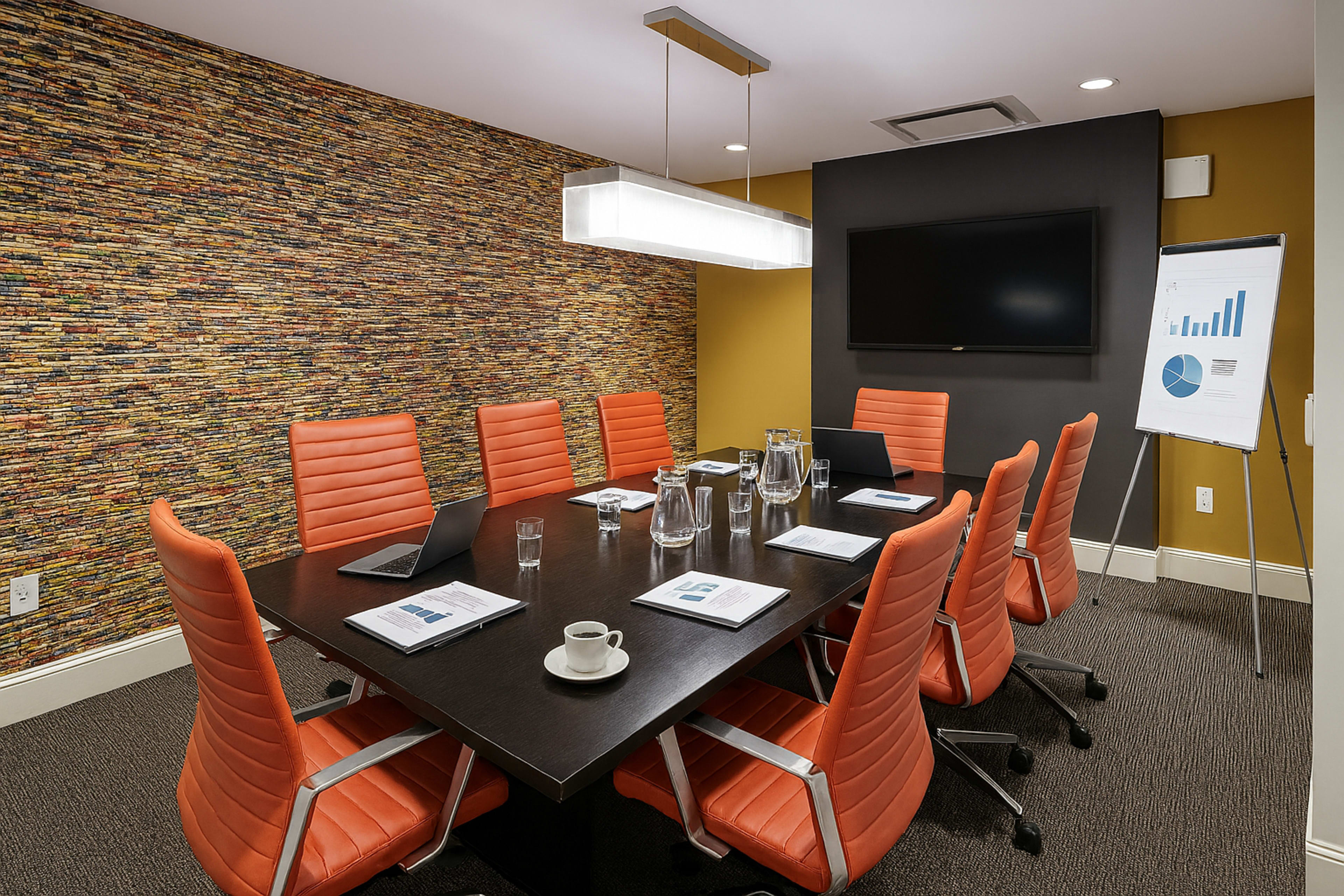 A modern conference room features a large table surrounded by eight orange chairs, a wall adorned with textured material, and a television displaying a presentation in the background.