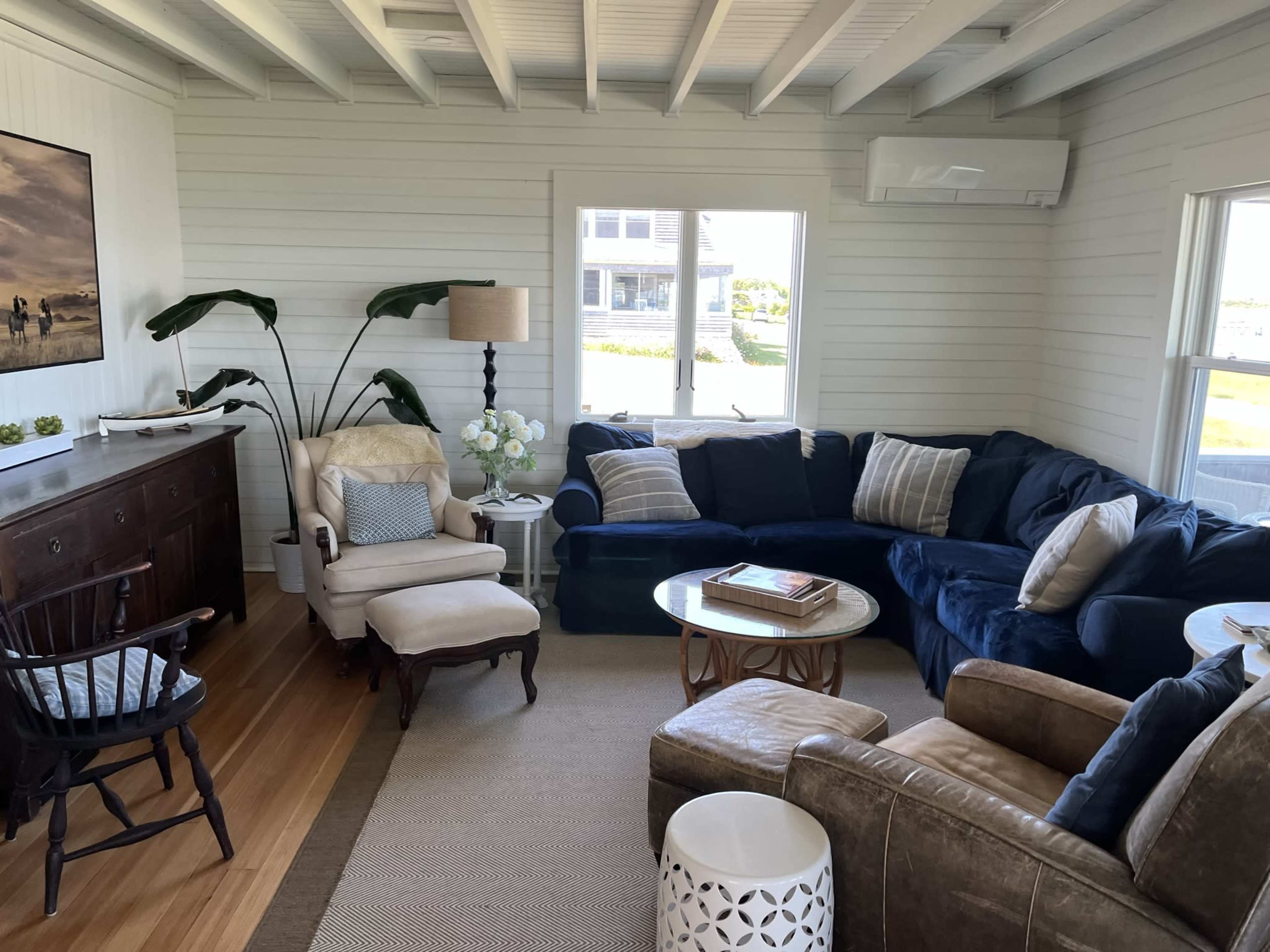 A bright living room with a mix of seating arrangements, including a navy blue sectional sofa, an armchair, and a coffee table, all arranged around a light-colored rug and wooden floors.