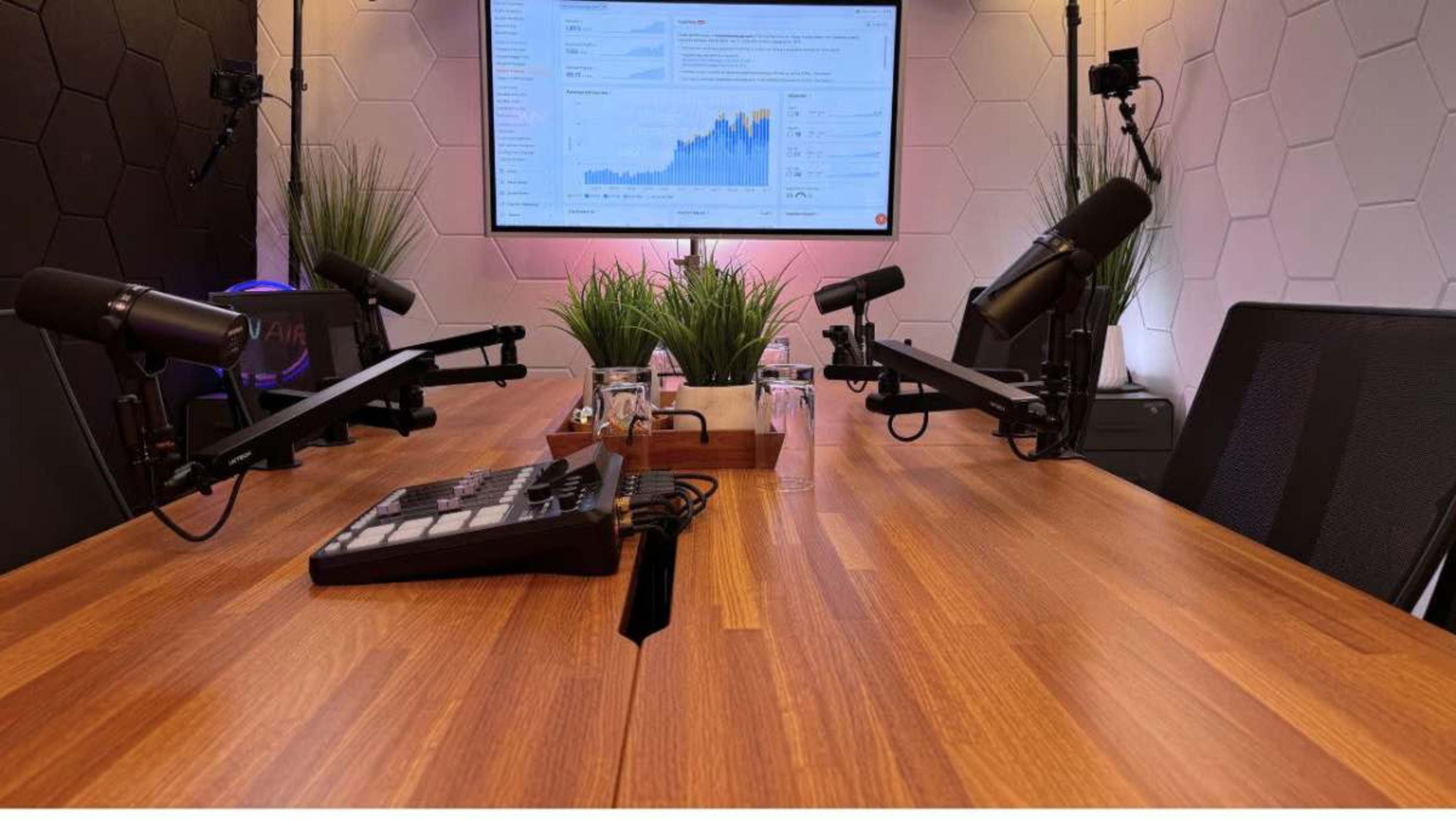 The image shows a modern podcast studio setup with a large screen displaying data analytics, two mounted microphones, and a wooden table with glass water cups.